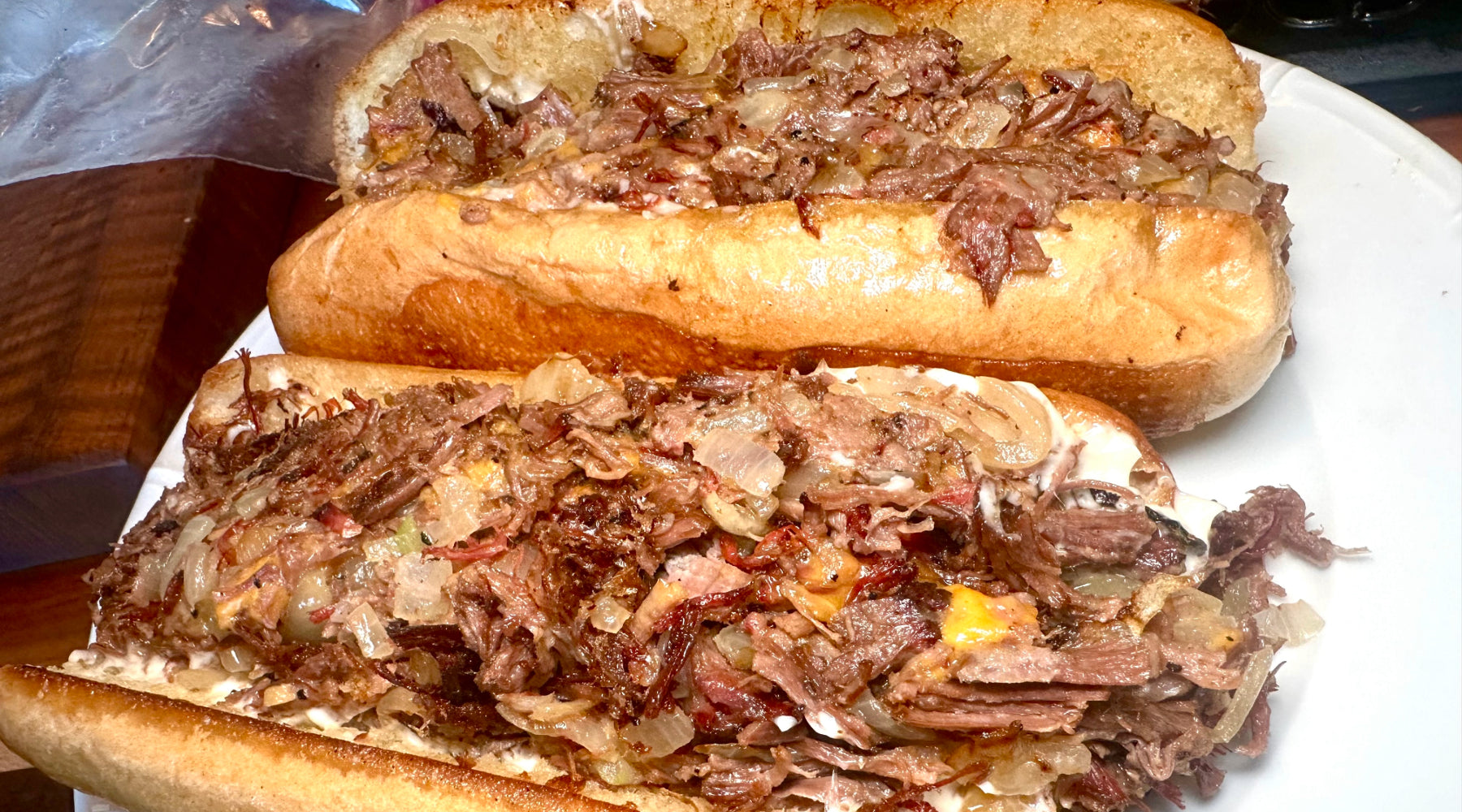 Two toasted hoagie rolls filled with brisket, caramelized onions, and melted cheese on a white plate.