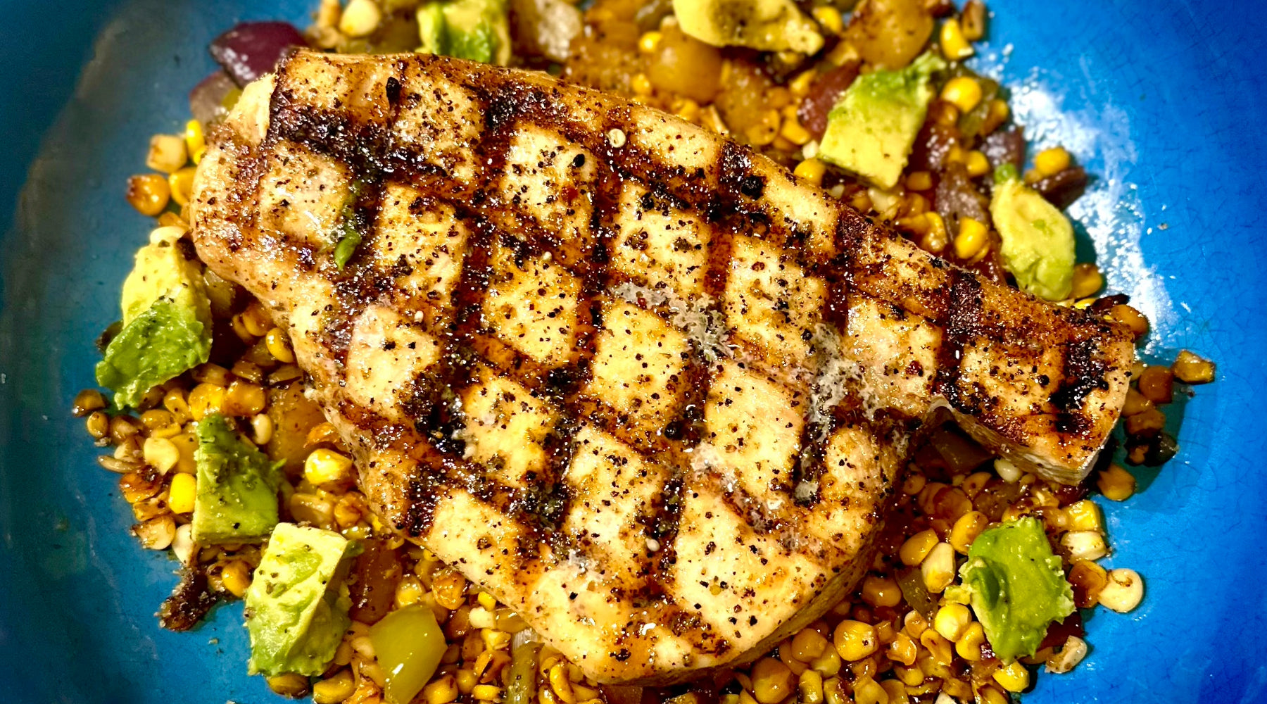 Grilled swordfish with seared grill marks over a bed of corn, avocado, and roasted vegetables on a blue plate.