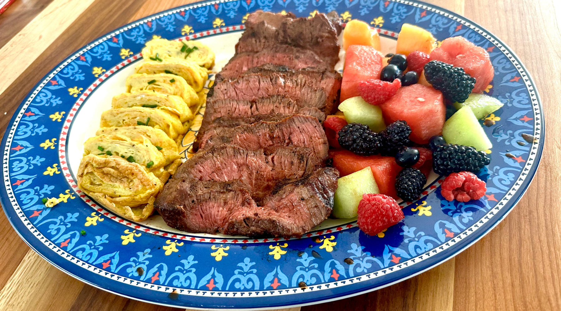 Sliced medium-rare steak with rolled omelet and fresh fruit, served on a decorative blue plate.