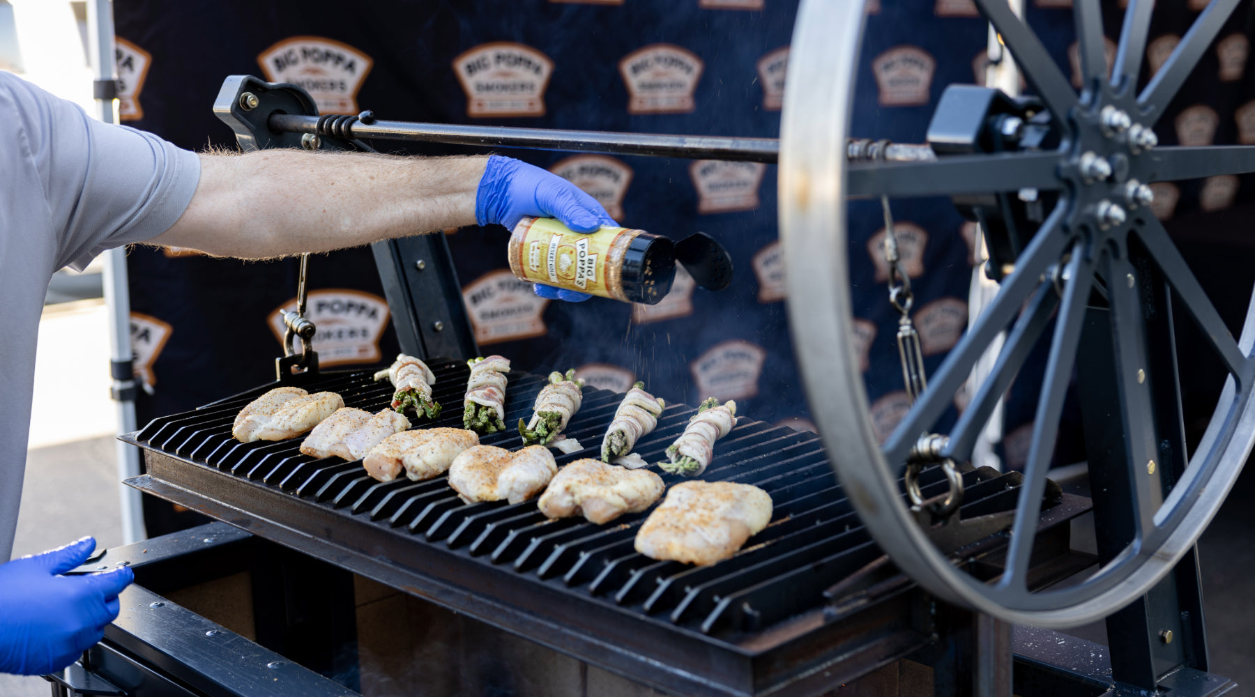 A person wearing blue gloves sprinkles seasoning on chicken and bacon-wrapped asparagus grilling over an open flame on a large smoker.