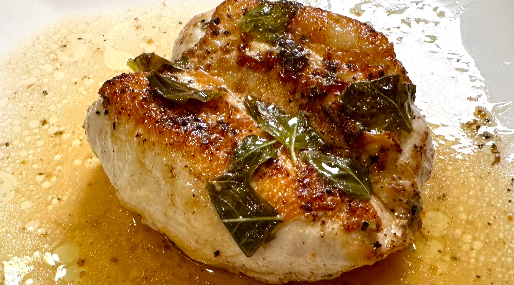 A close-up of a golden-brown seared chicken breast topped with fresh herbs, glistening with pan juices on a white plate.