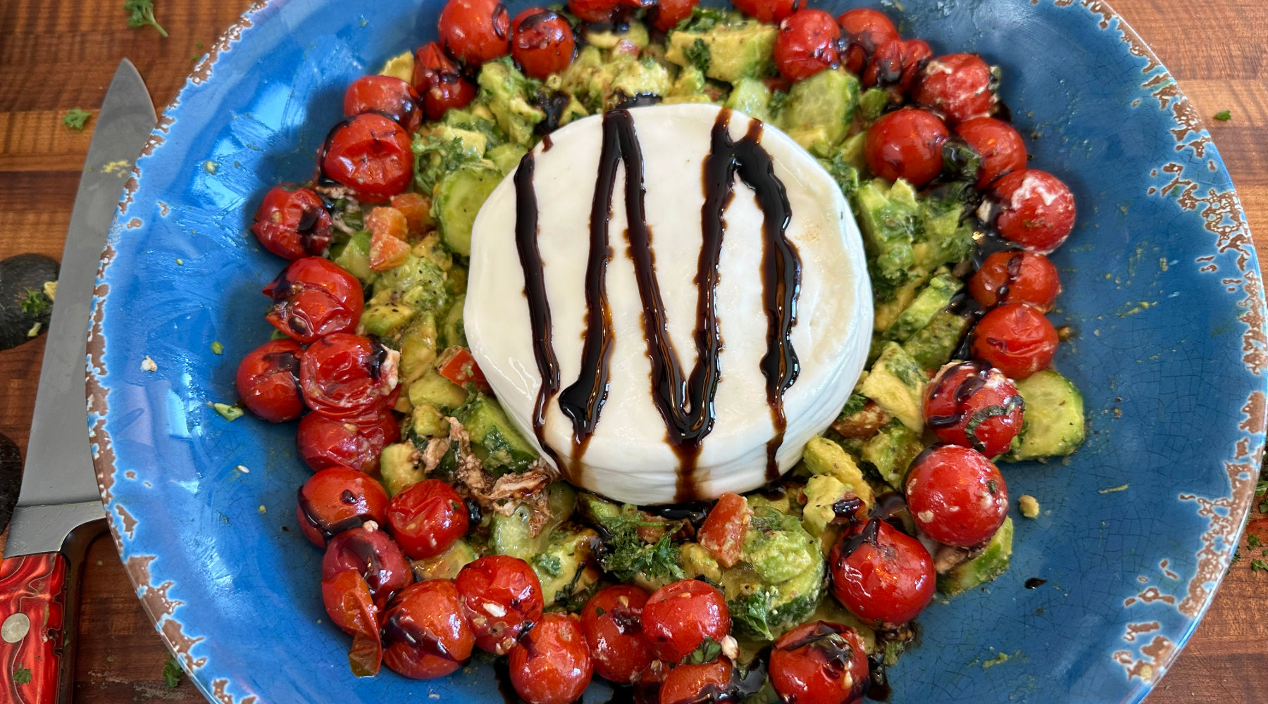 A blue plate with creamy burrata drizzled with balsamic glaze, surrounded by roasted cherry tomatoes and avocado salad.