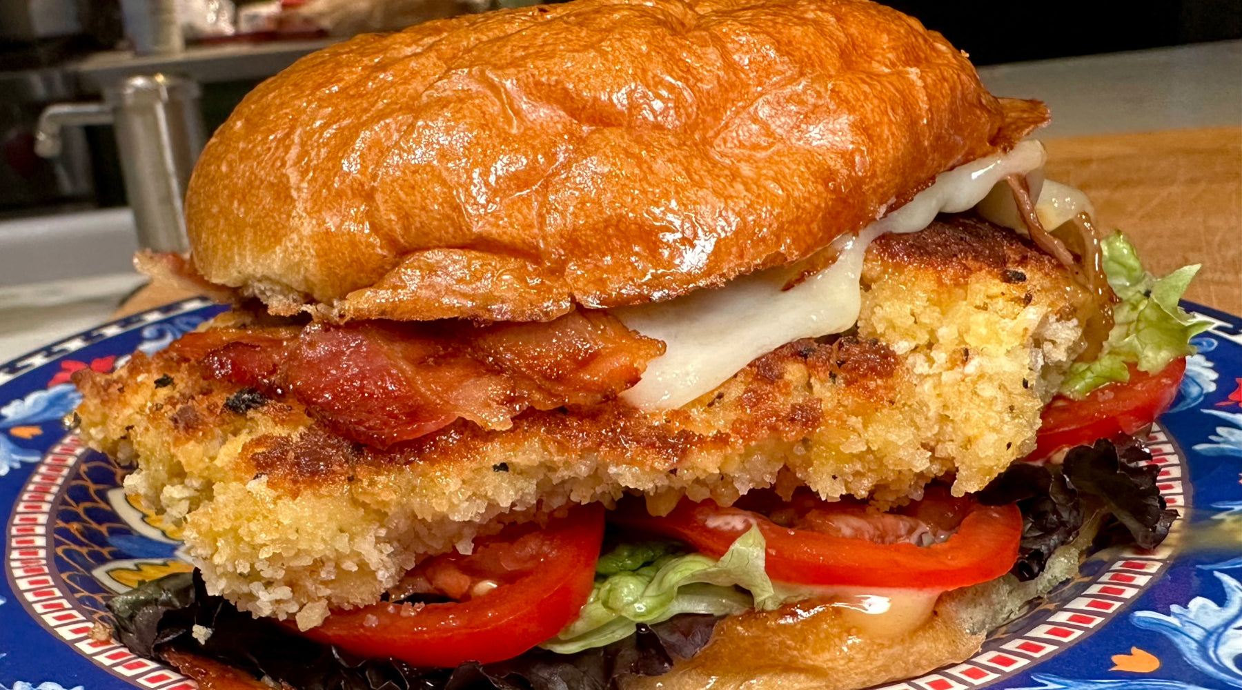 Crispy fried chicken sandwich with bacon, melted cheese, lettuce, and tomato on a toasted bun, served on a decorative plate.