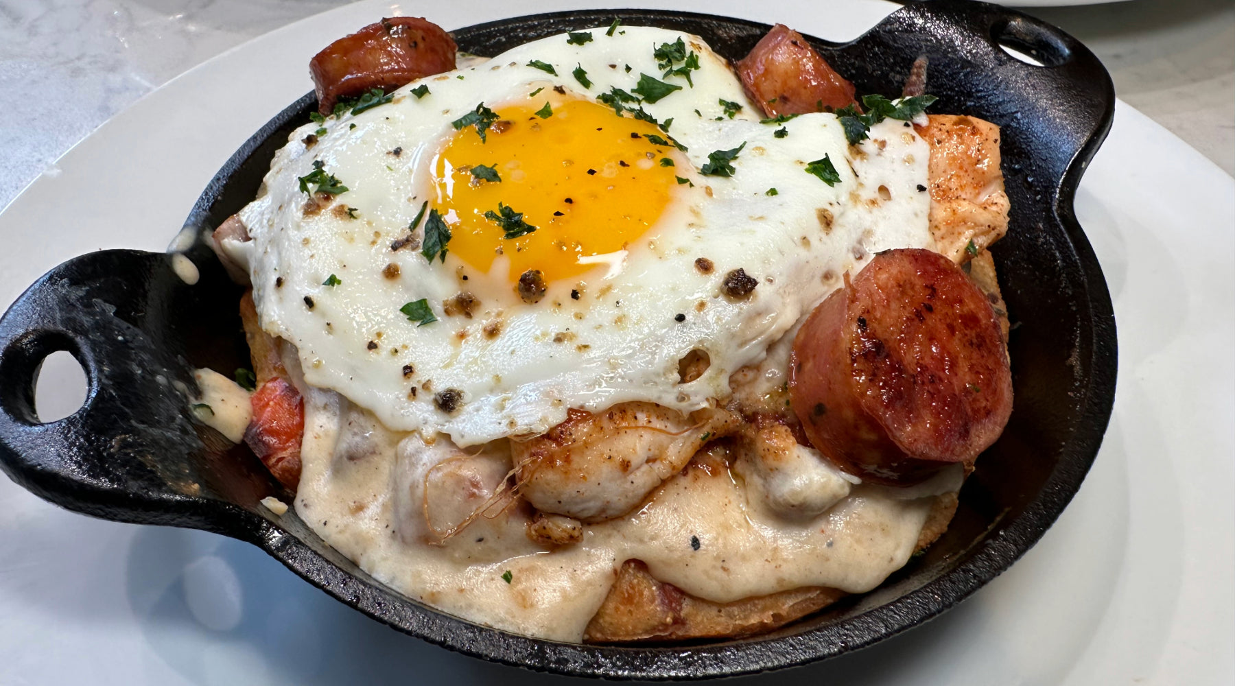 A cast iron skillet with a cheesy dish topped with a sunny-side-up egg, grilled sausage, and fresh herbs, served on a white plate.