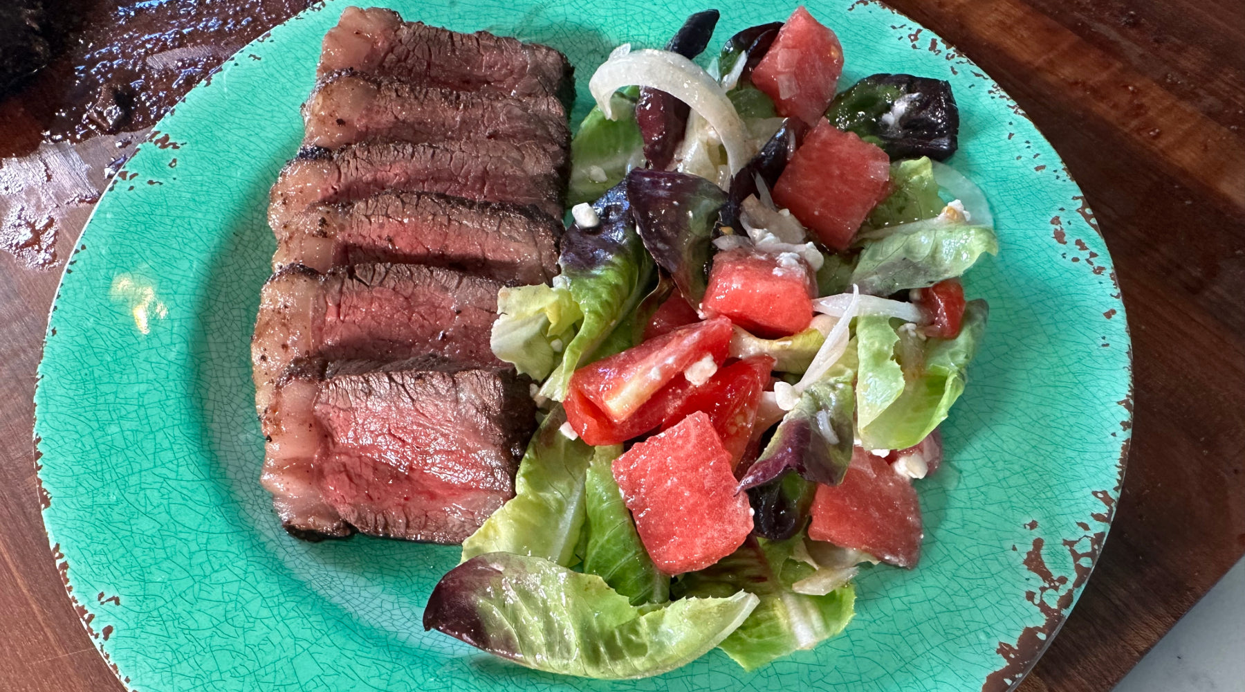 Sliced medium-rare steak with a fresh salad of mixed greens, watermelon, tomatoes, onions, and feta, served on a turquoise plate.