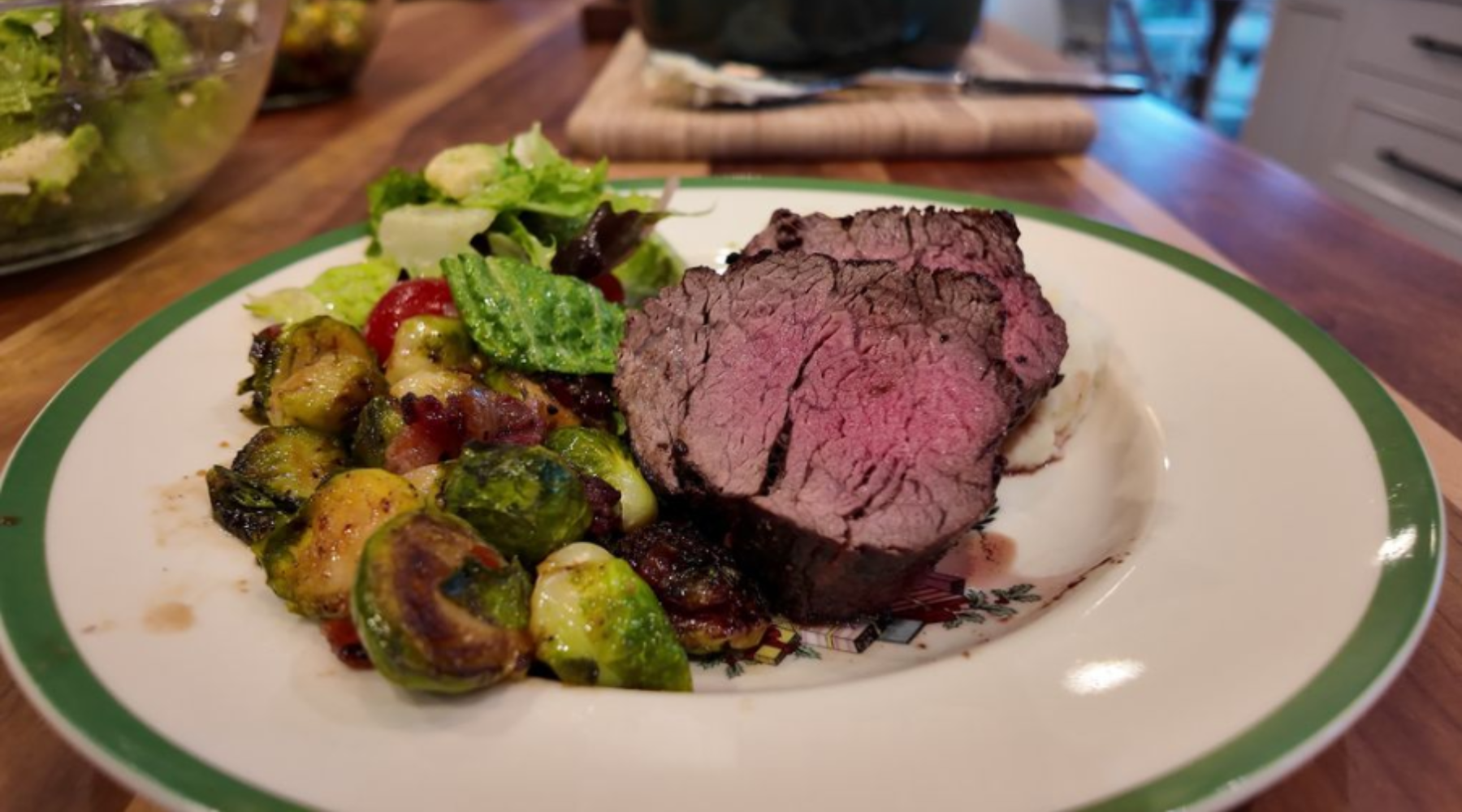 Sliced medium-rare beef tenderloin with roasted Brussels sprouts, bacon, and a fresh salad, served on a white plate with a green rim.