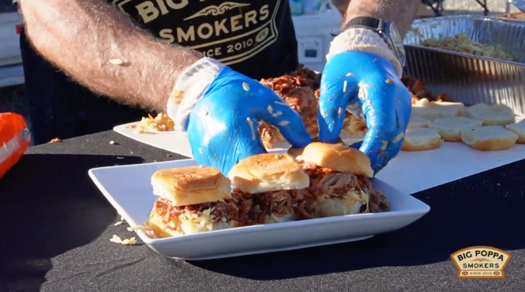 A person wearing blue gloves assembles pulled pork sliders on a white plate at an outdoor event.