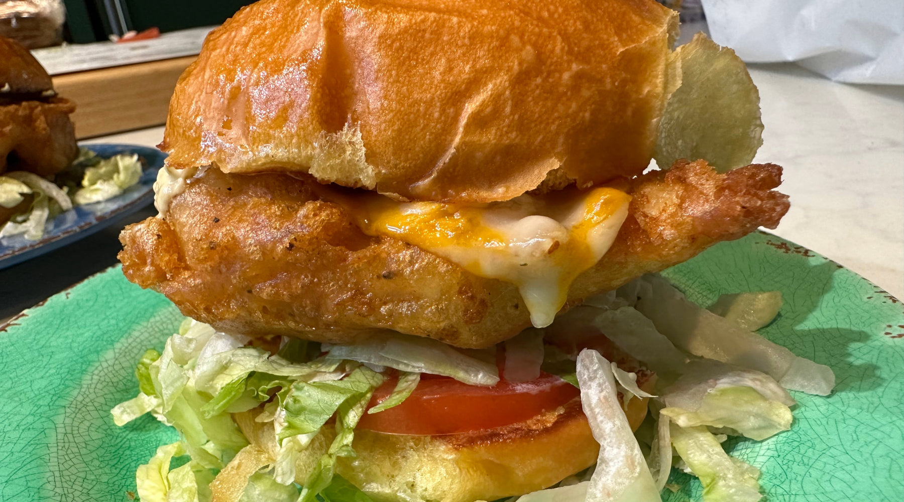 Crispy fried fish sandwich with melted cheese, lettuce, and tomato on a toasted bun, served on a green plate.