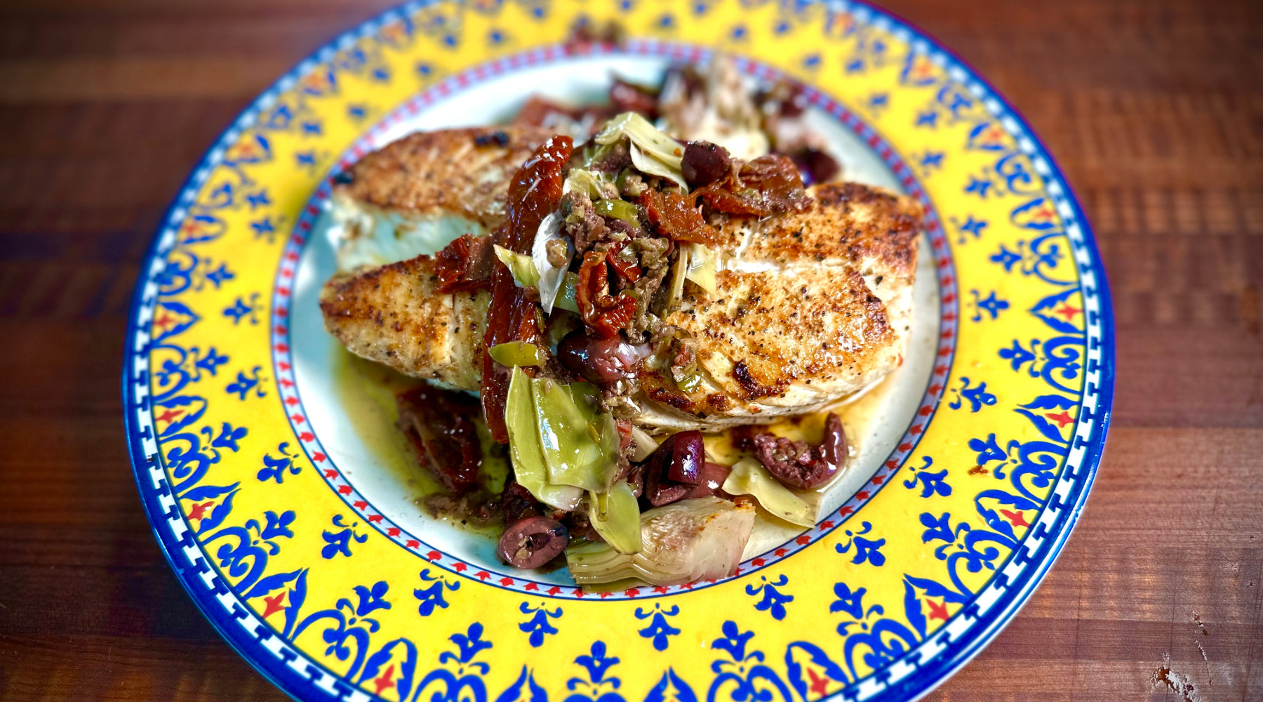 Grilled halibut topped with sun-dried tomatoes, olives, and artichokes, served on a colorful patterned plate.