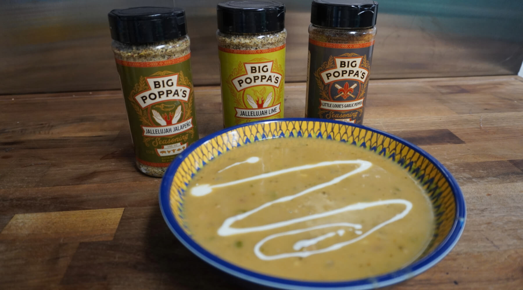 Bowl of creamy soup with a swirl of white sauce, placed in front of three Big Poppa’s seasoning bottles on a wooden counter.
