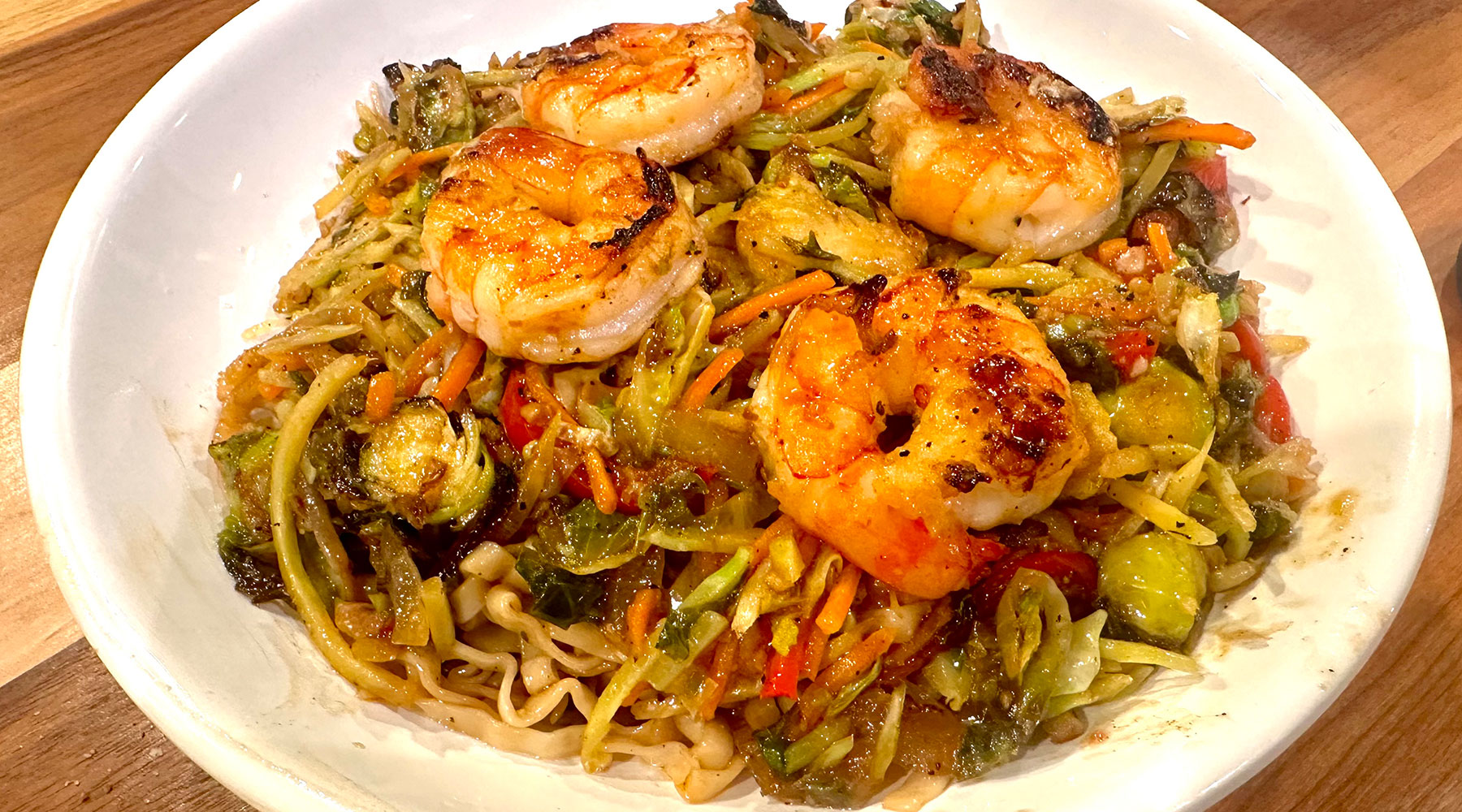 A hearty meal of grilled shrimp and stir-fried vegetables served in a white bowl. The vegetables, including Brussels sprouts and carrots, are mixed with noodles and seasoned to a golden brown. The dish is presented on a wooden tabletop.