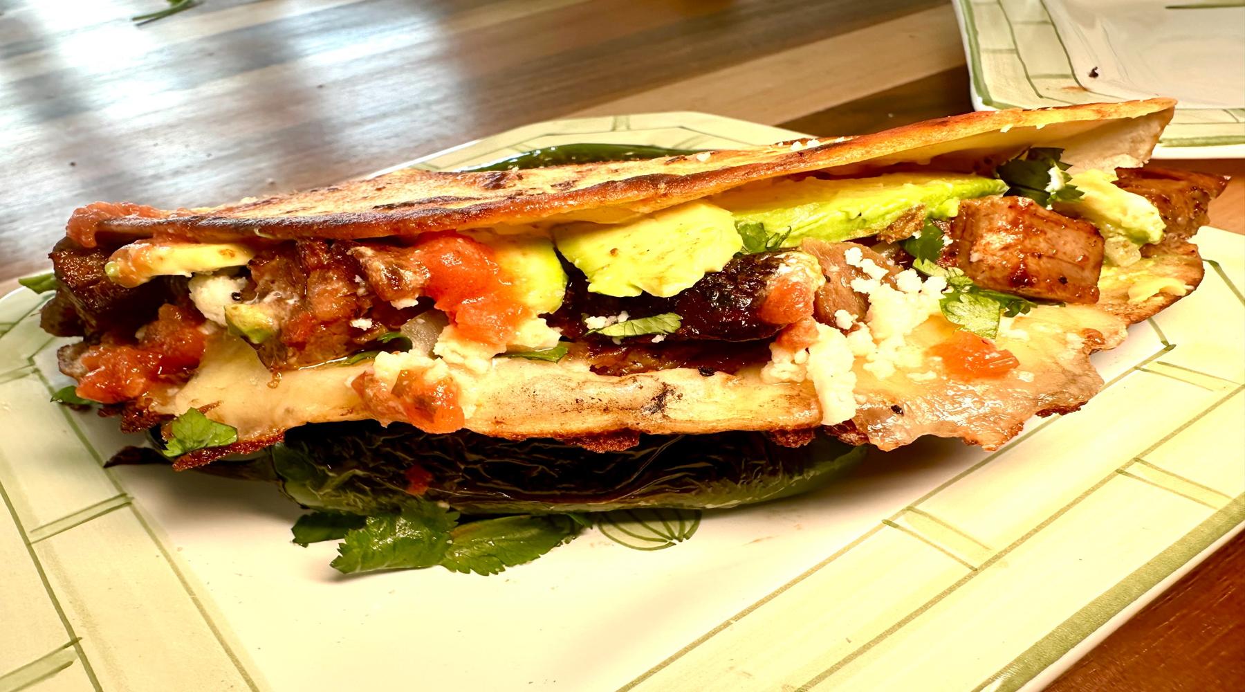 A close-up of a brisket quesataco filled with chunks of tender brisket, melted cheese, avocado slices, and crumbled cheese, served with a grilled jalapeño on the side.