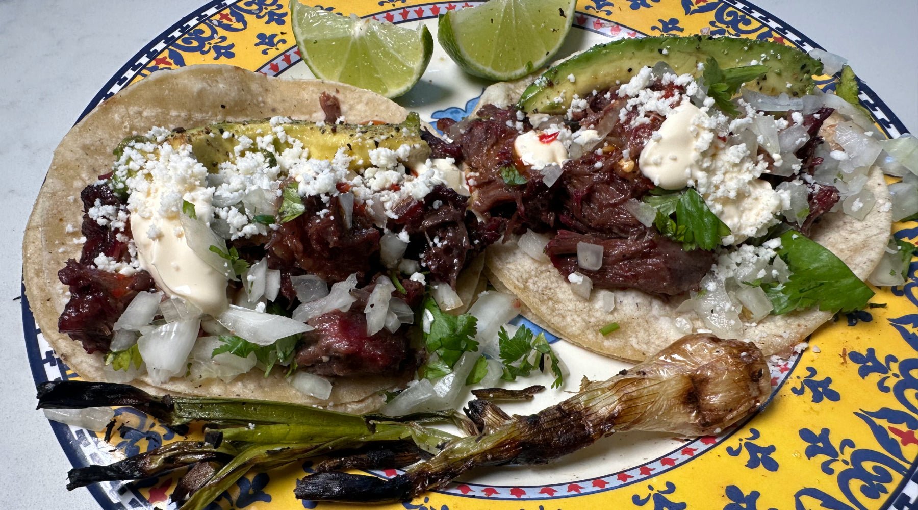 Two soft corn tacos filled with tender, shredded beef, topped with diced onions, fresh cilantro, crumbled white cheese, avocado slices, and a dollop of crema, served on a decorative plate with a charred green onion and lime wedges on the side.