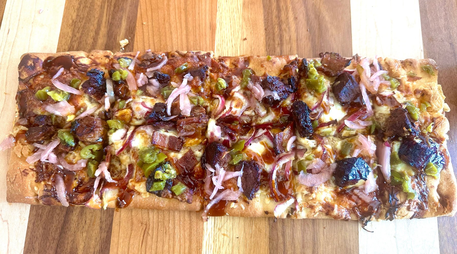 A flatbread pizza cut into squares, featuring toppings such as barbecued meat, pickled green peppers, red onions, and barbecue sauce, placed on a wooden board.