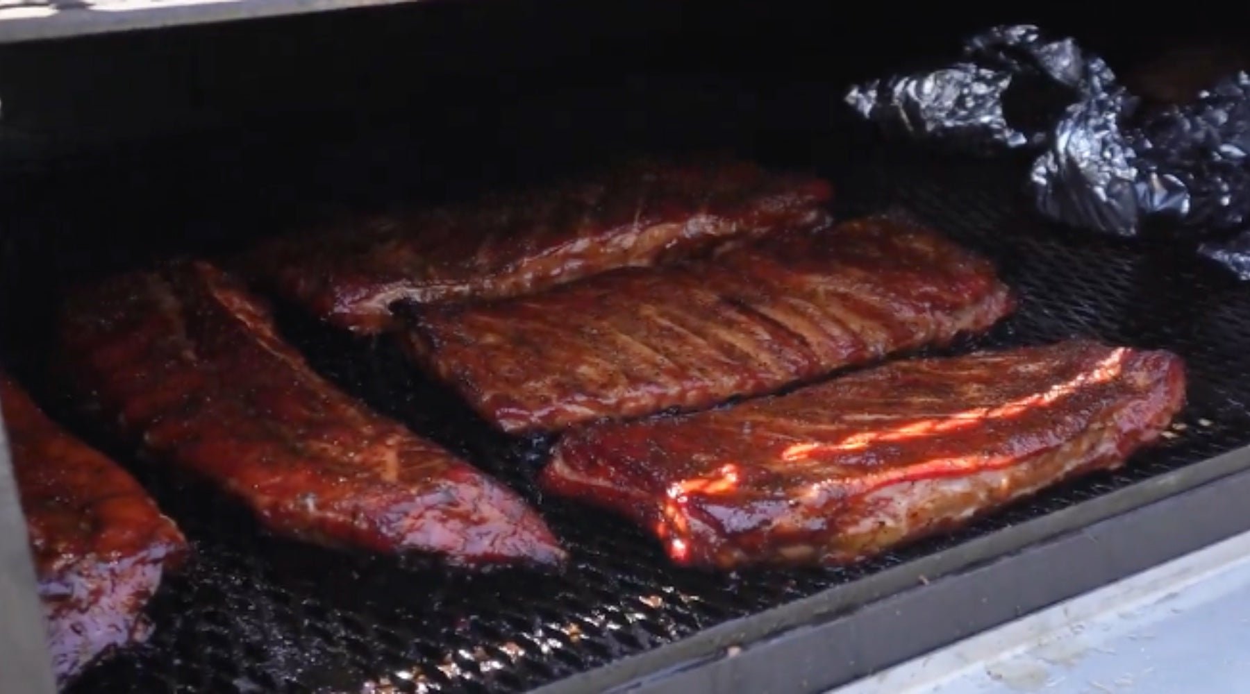 Several racks of pork ribs coated in a rich, reddish-brown glaze are being smoked on a grill, with a light sheen indicating they are almost fully cooked. In the background, there are some foil-wrapped items also being smoked.