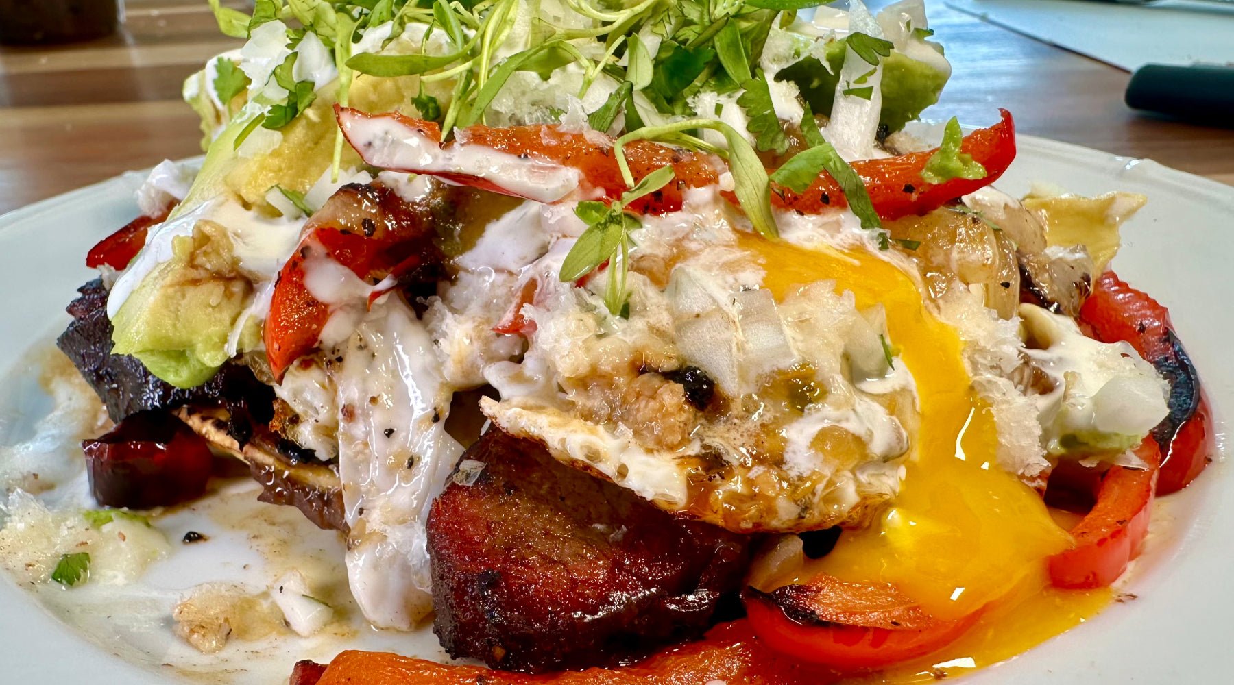 A colorful dish featuring a perfectly fried egg with a runny yolk, topped with red bell peppers, sliced avocado, and fresh greens, all drizzled with a creamy sauce.