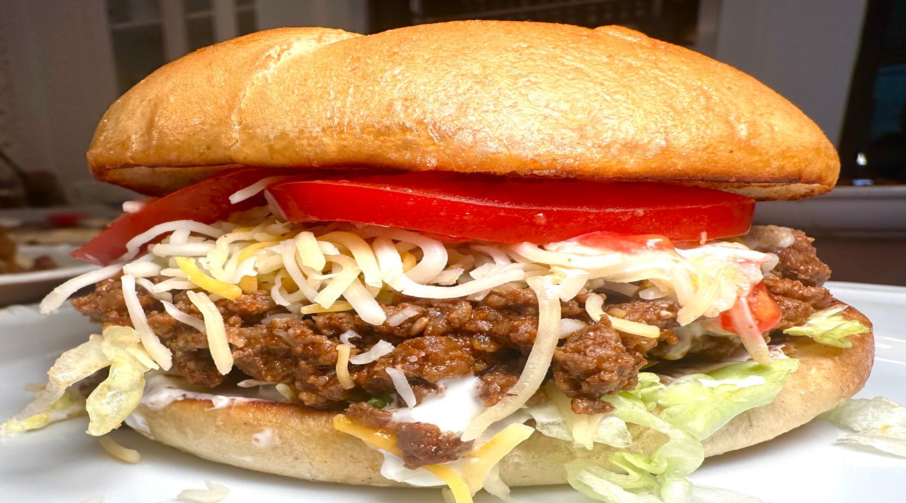 A hearty taco burger with seasoned ground beef, shredded cheese, fresh tomato slices, lettuce, and a soft bun, showcasing a delicious fusion of taco and burger.