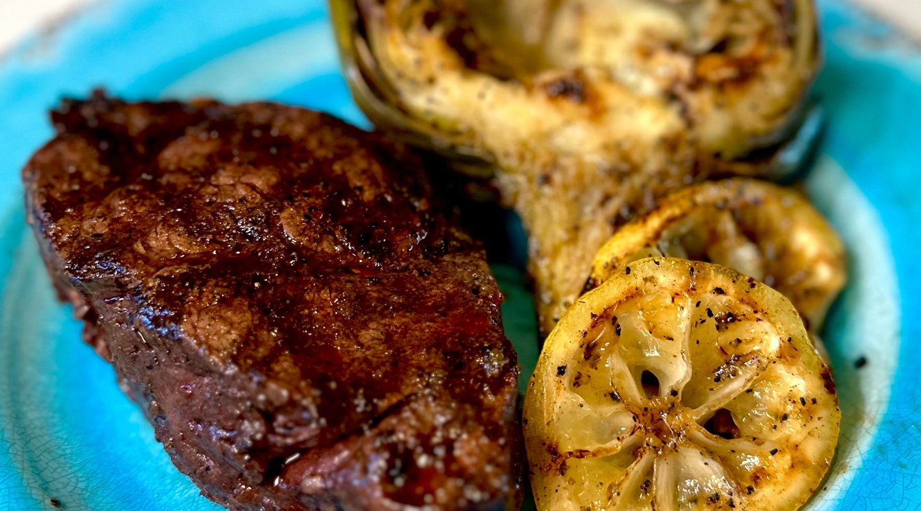 A grilled steak served on a blue plate alongside a roasted artichoke half and two slices of charred lemon. The steak has a dark sear, and the artichoke shows grill marks.