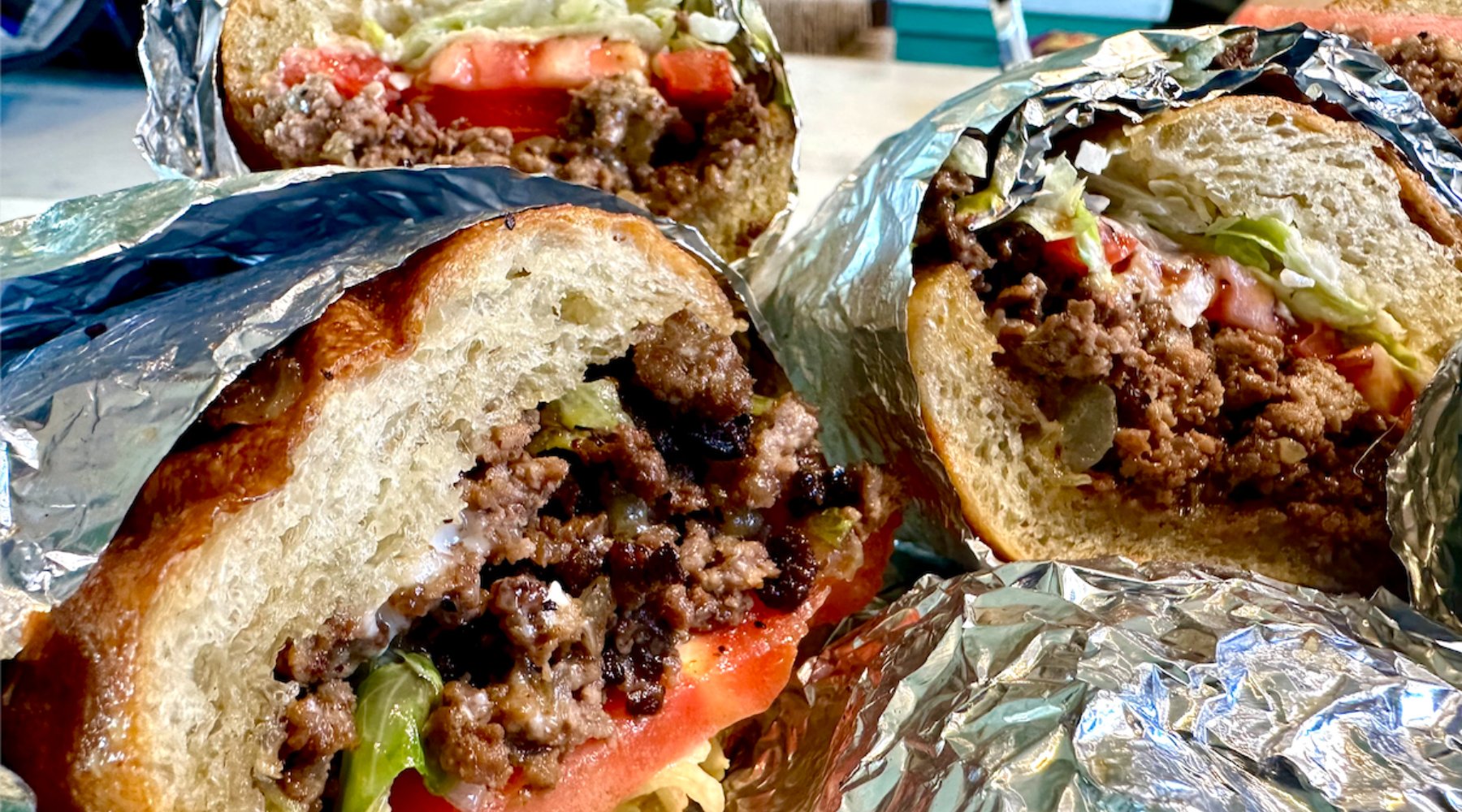 Close-up of three large sandwiches wrapped in foil, each cut in half to reveal ground beef, lettuce, and tomato inside crusty bread.