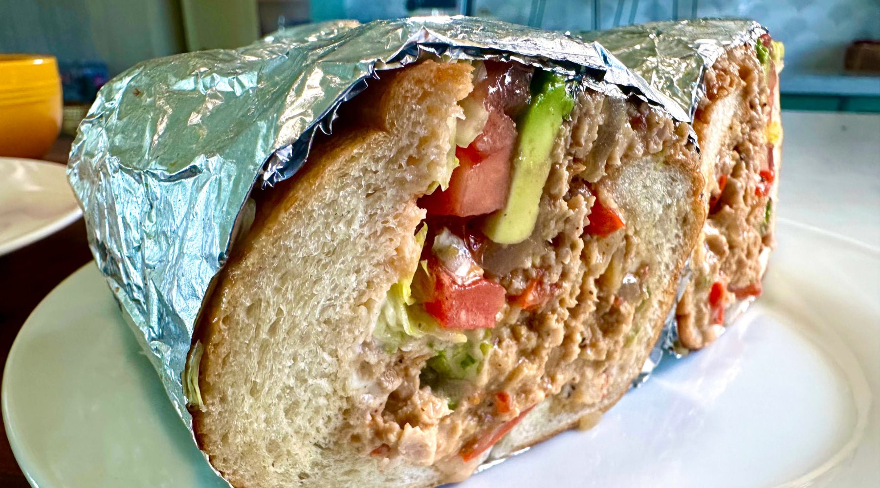 A large sandwich filled with chicken, avocado, tomato, and lettuce, wrapped in aluminum foil and served on a white plate. The sandwich is cut to show the thick, soft bread and generous filling.