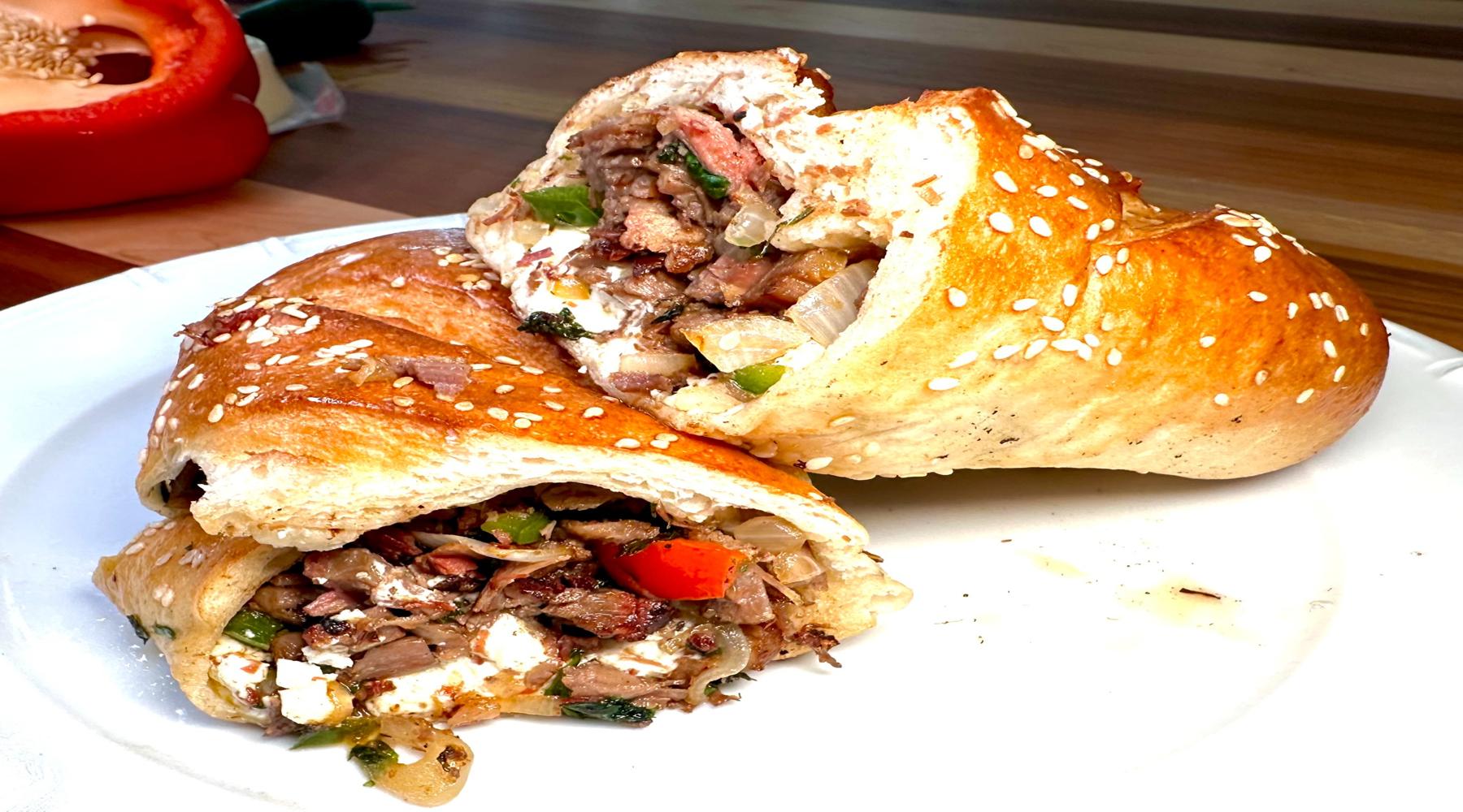 A sesame seed-topped sandwich cut in half, revealing a hearty filling of meat, onions, bell peppers, and cheese. The sandwich sits on a white plate, highlighting its golden-brown crust.