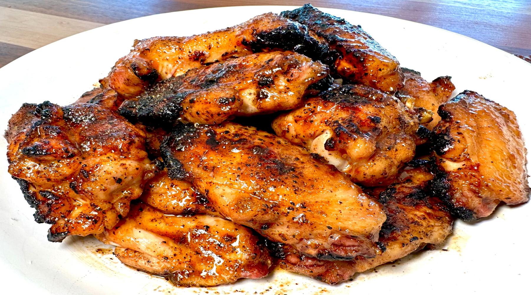 A plate of honey sriracha chicken wings, charred to a golden brown with a glossy, sticky glaze. The wings are seasoned and grilled, showcasing a mix of crispy and tender textures.