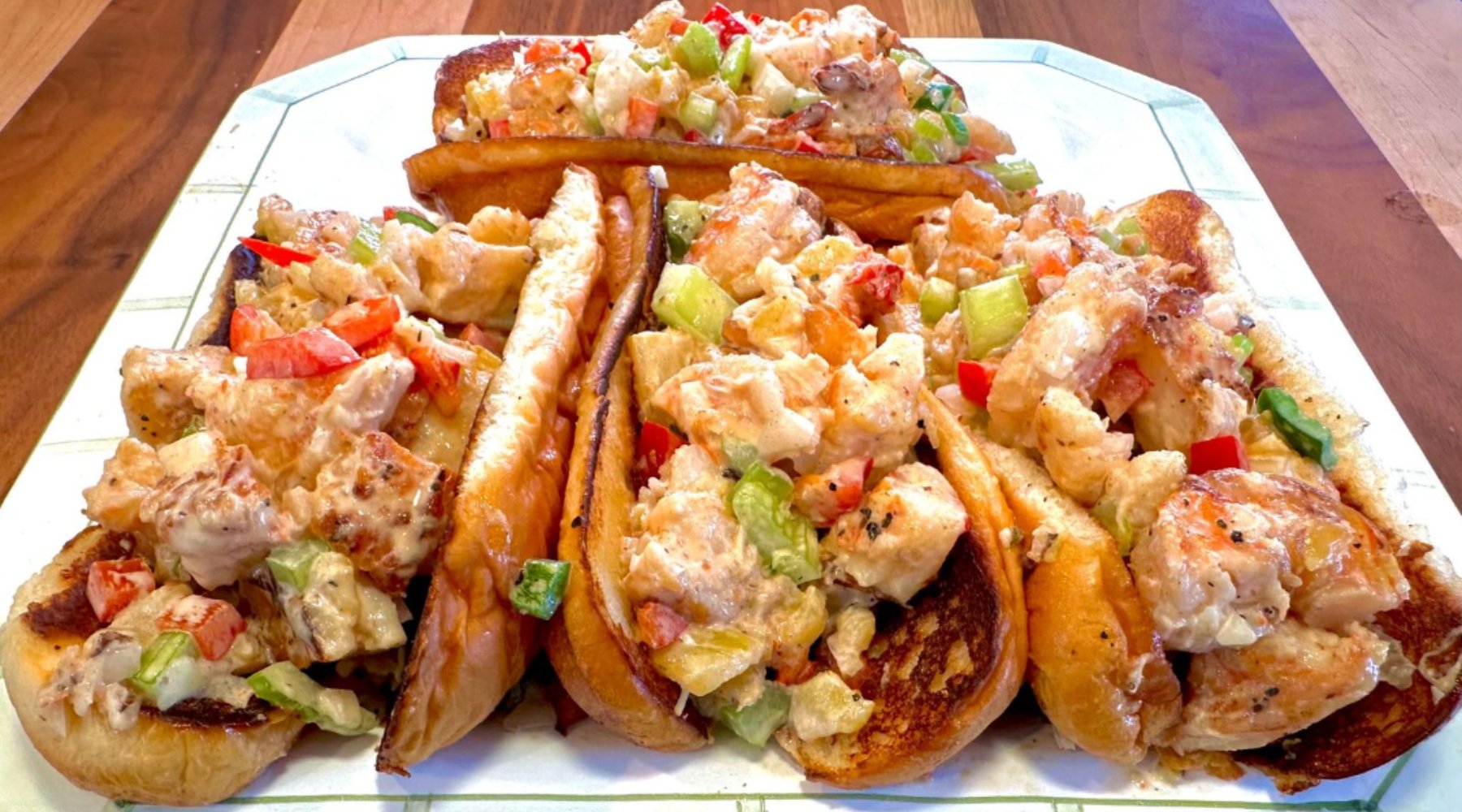 A platter of shrimp rolls filled with chunks of shrimp, diced celery, and red bell peppers, all mixed in a creamy dressing, served in toasted buns.