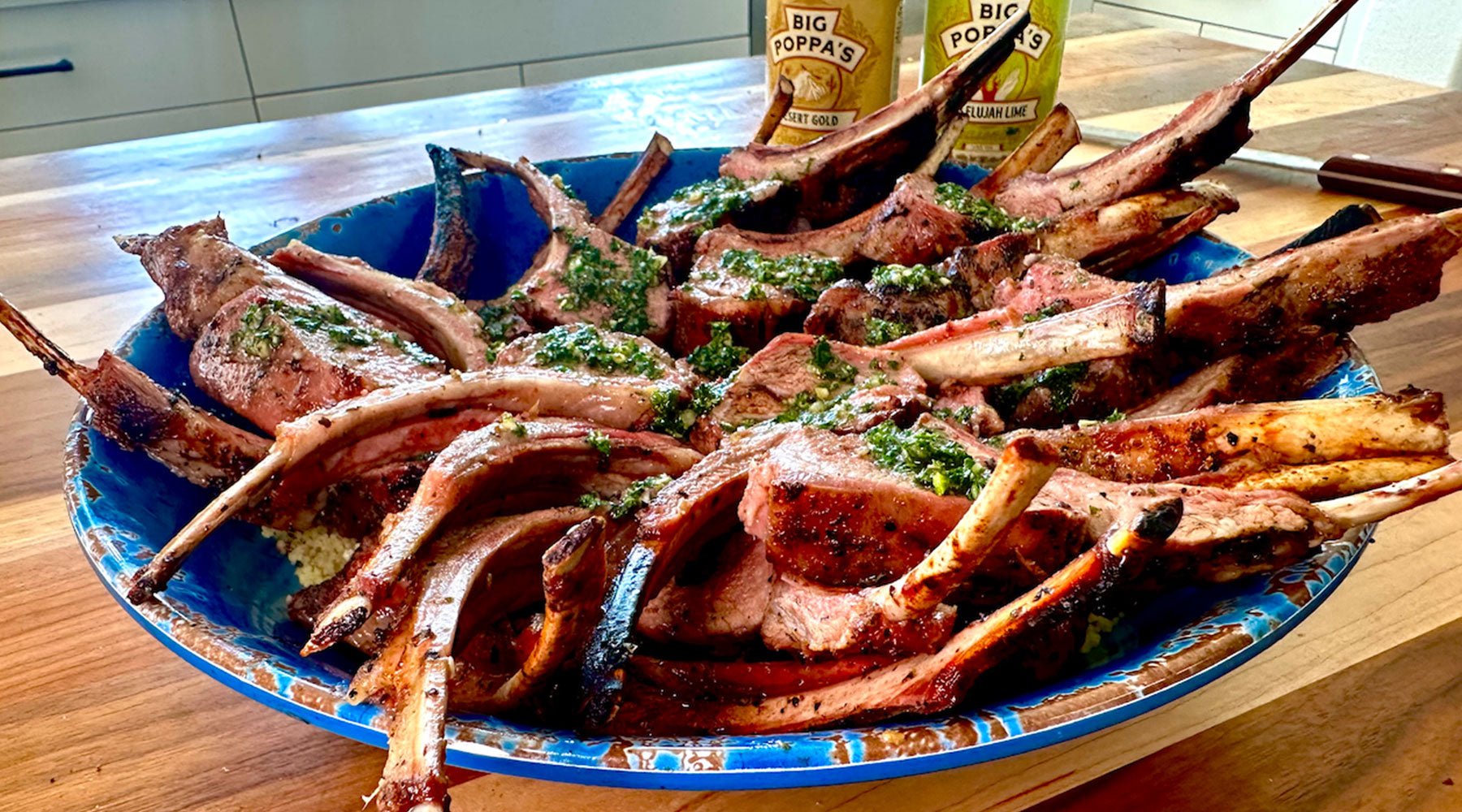 A large blue platter filled with grilled lamb chops, garnished with a green herb sauce, on a wooden countertop with seasoning containers in the background.