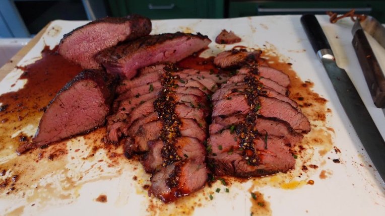 A sliced piece of medium-rare tri-tip displayed on a white cutting board, with a seared crust and a juicy pink center, drizzled with sauce and garnished with chili crunch.