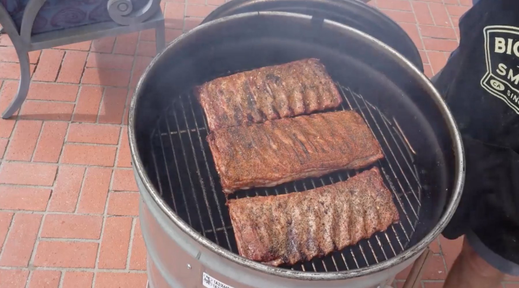 Three slabs of seasoned ribs being smoked on a cylindrical smoker, with smoke rising from the grill, set on a brick patio with a person in the background wearing a black apron.