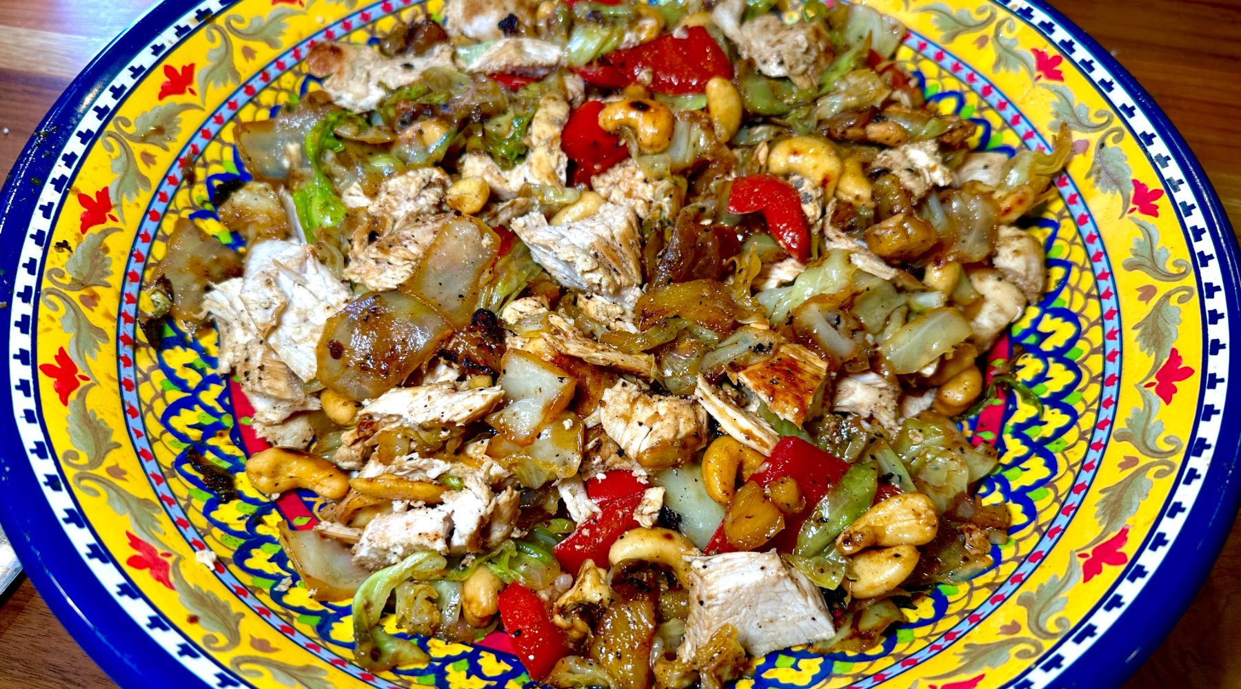 A colorful dish served in a vibrant, ornate ceramic bowl. The dish consists of sliced grilled chicken, cashews, red bell peppers, onions, and sautéed vegetables, all seasoned and mixed together.