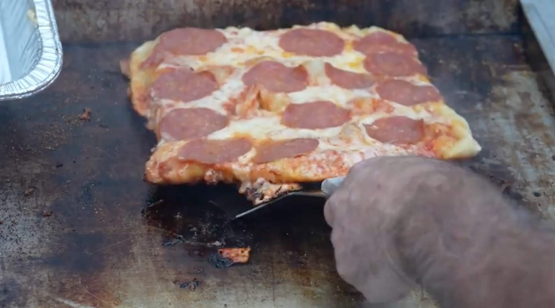 A close-up of a large rectangular pepperoni pizza being cooked on a flat grill. A person's hand is using a spatula to lift the pizza, showing melted cheese and pepperoni slices on top.