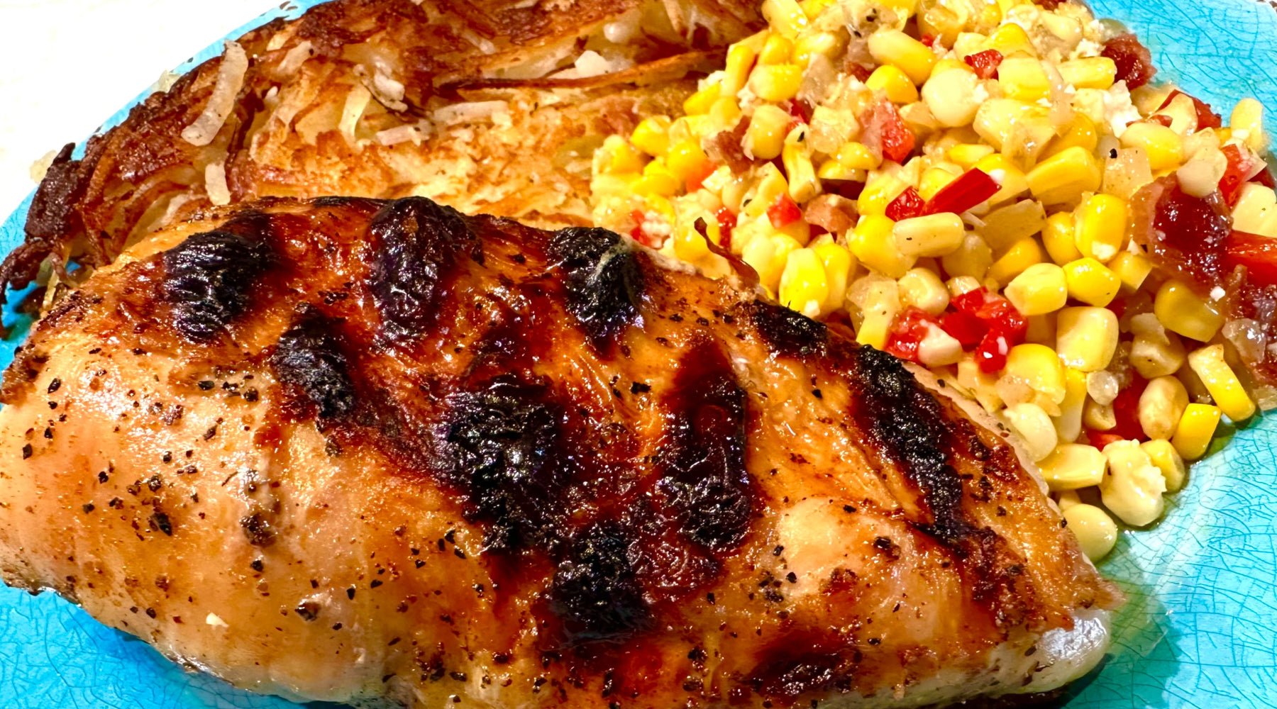 A close-up of a grilled chicken breast with a charred surface, served with a side of crispy hash browns and a colorful corn salad with red bell peppers on a blue plate.