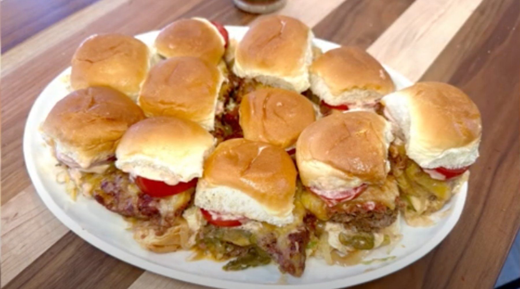 A platter of small sliders with soft, golden buns, each containing a beef patty, melted cheese, tomato slice, and other toppings, arranged on a white plate.