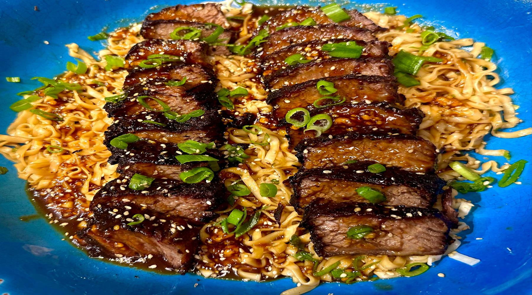 A close-up of a dish featuring sliced barbecued beef short ribs served over a bed of noodles in a blue bowl. The ribs are glazed with a rich sauce and sprinkled with sesame seeds and chopped green onions.