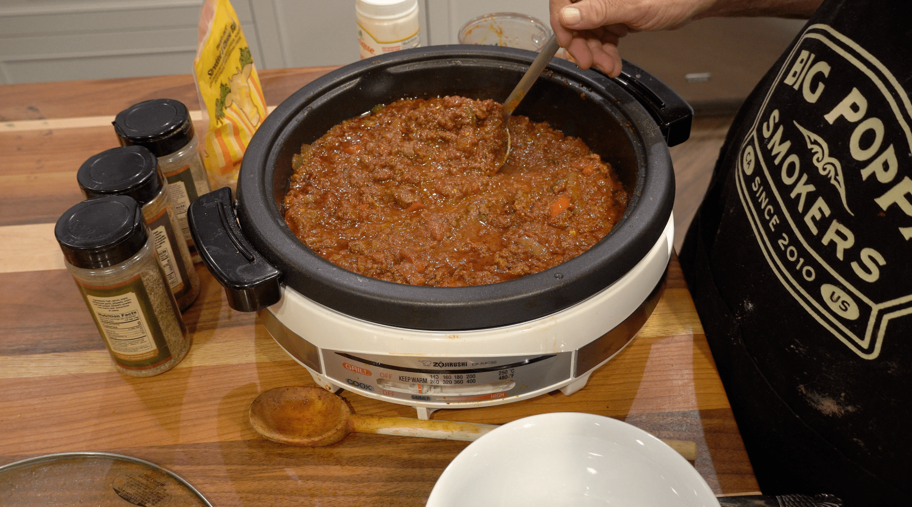 A slow cooker filled with a rich, chunky meat and tomato sauce being stirred with a ladle, surrounded by seasonings and kitchen tools.