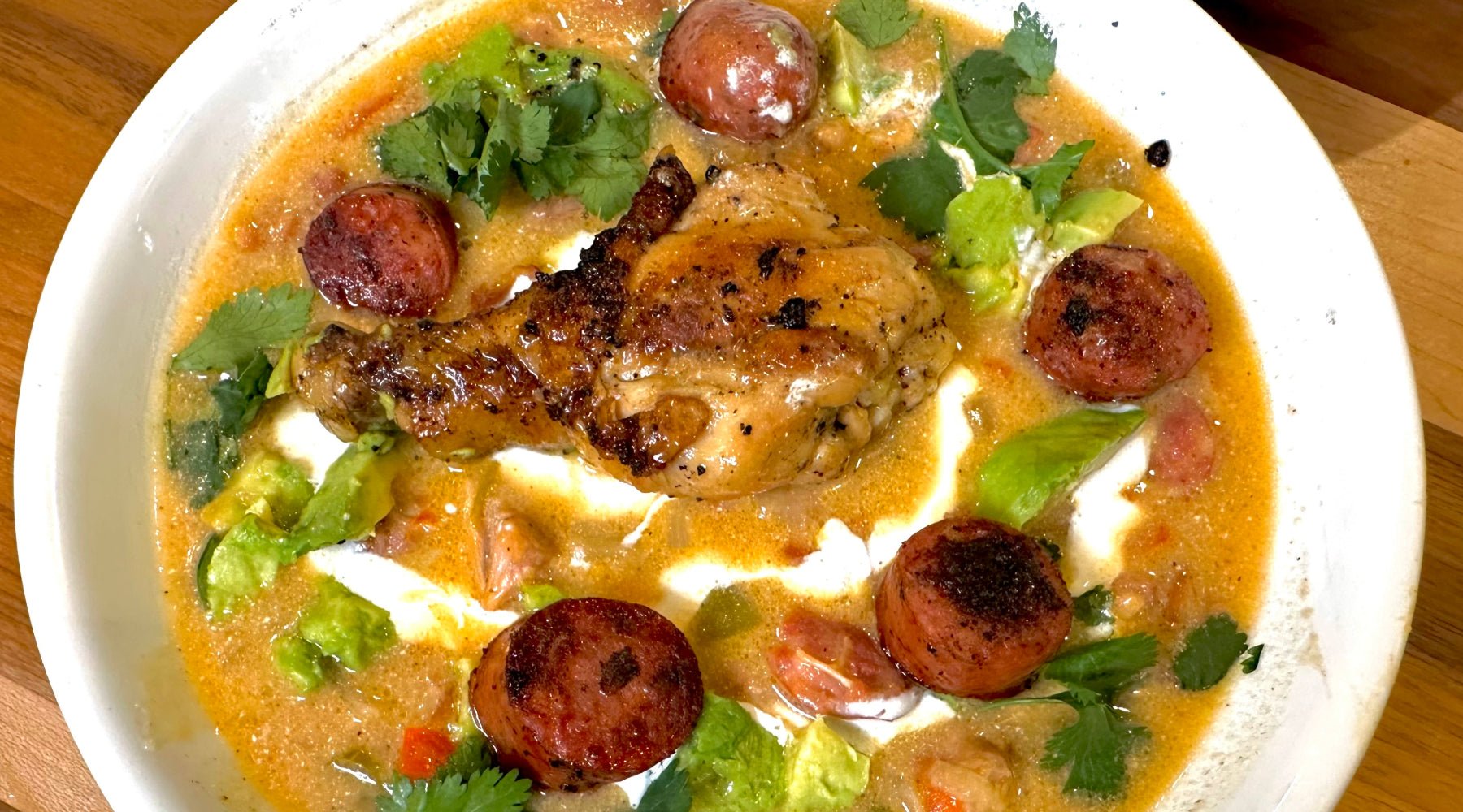 A bowl of soup featuring a roasted chicken leg, seared sausage slices, avocado chunks, cilantro, and a creamy broth with tomatoes.