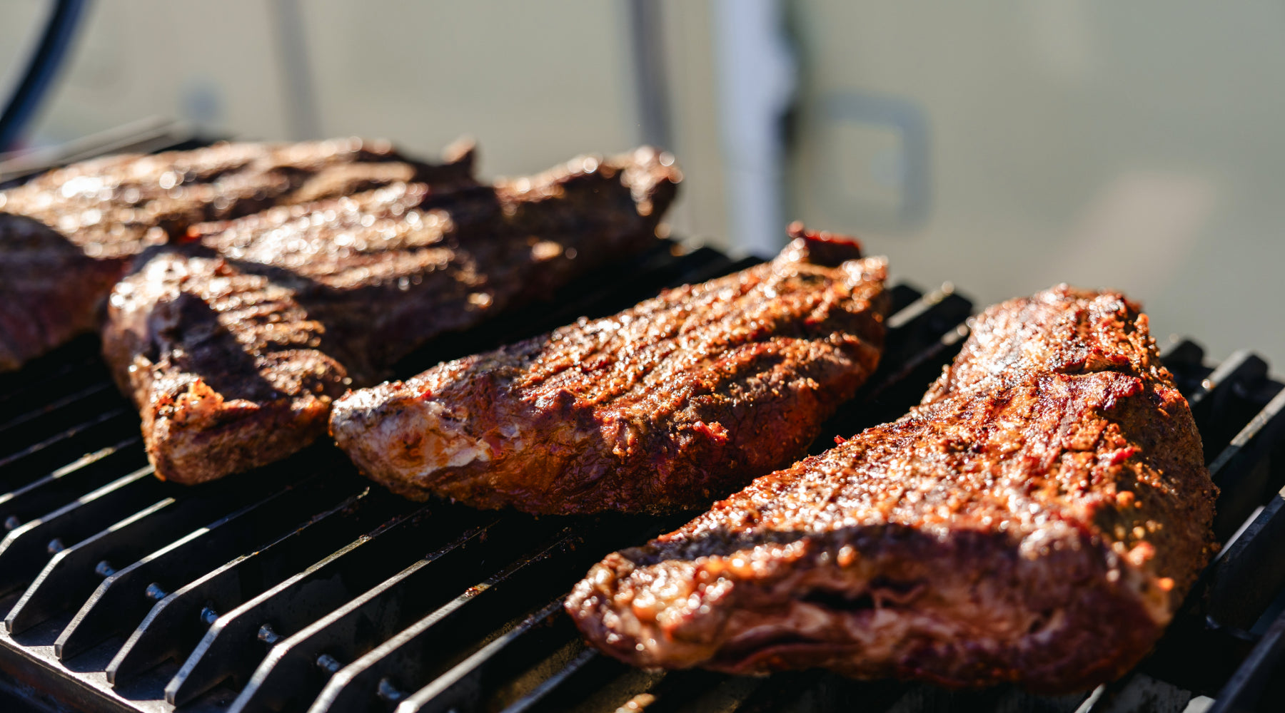 Several large cuts of marinated steak cooking on a grill, with grill marks visible on the meat, set against an outdoor background.