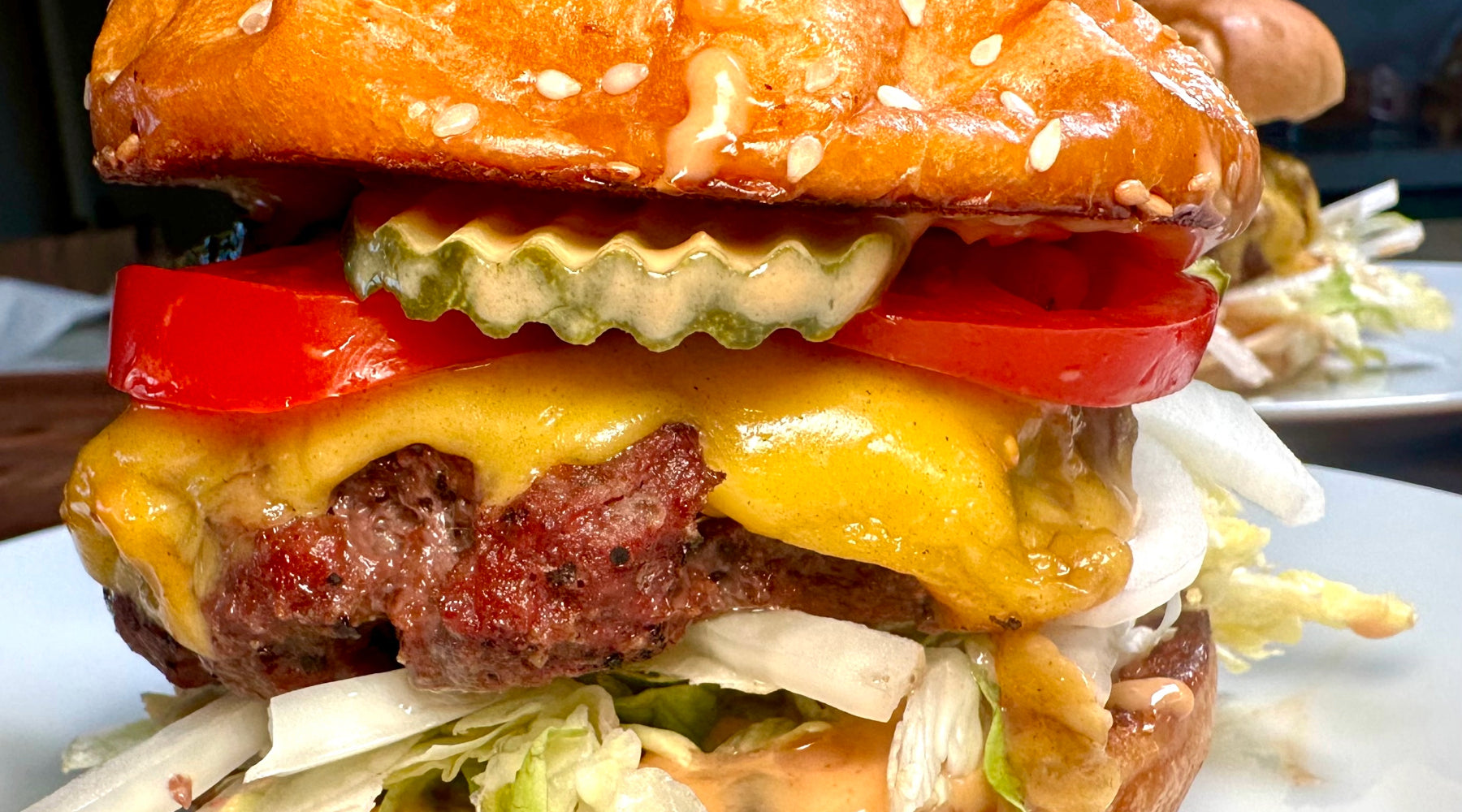 A close-up of a juicy cheeseburger with a sesame seed bun, topped with melted cheese, thick tomato slices, crinkle-cut pickles, and shredded lettuce, with a creamy sauce dripping down.