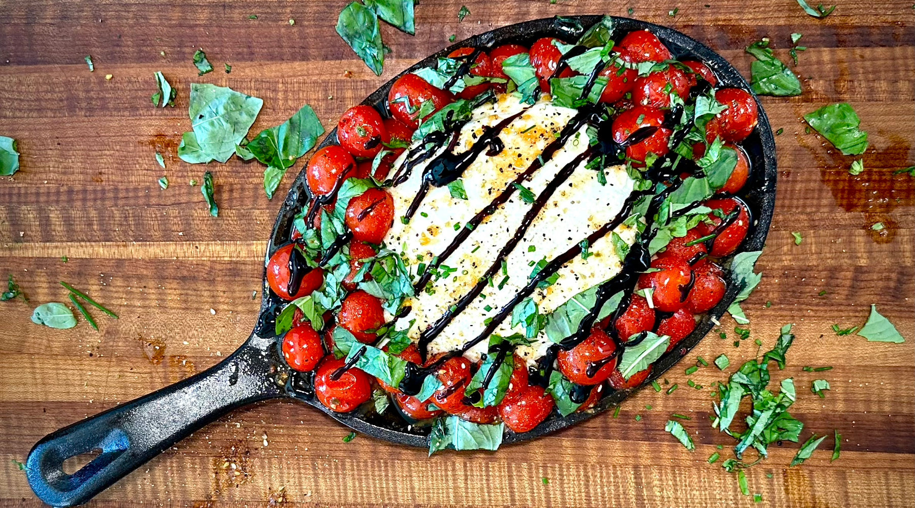 A cast iron skillet on a wooden surface contains a dish of roasted cherry tomatoes and fresh basil leaves, surrounding a large piece of burrata cheese drizzled with balsamic glaze.