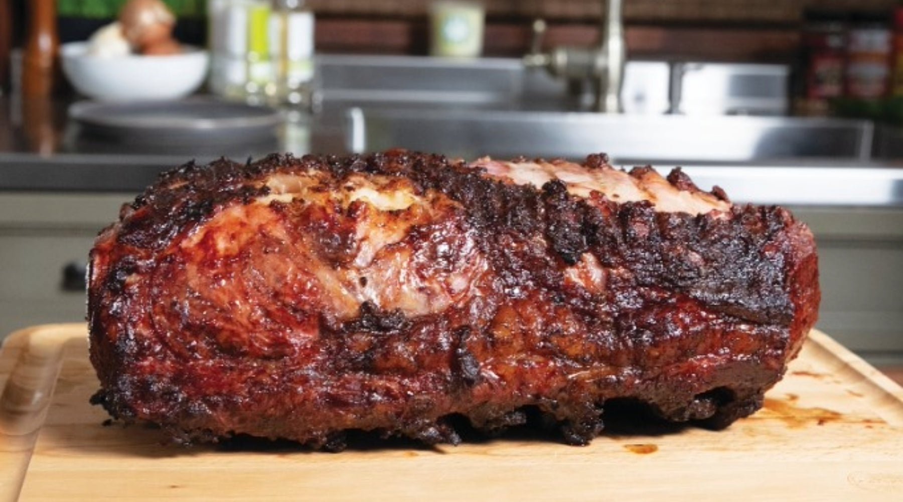 A perfectly cooked prime rib roast with a crispy, golden-brown crust and juicy, marbled interior resting on a wooden cutting board in a kitchen setting.