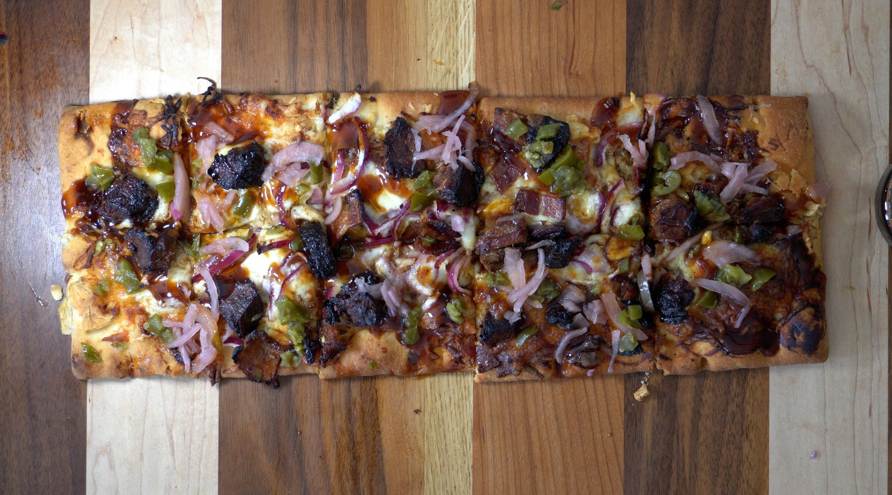 Rectangular BBQ brisket pizza topped with cheese, jalapeños, red onions, and barbecue sauce on a wooden board.