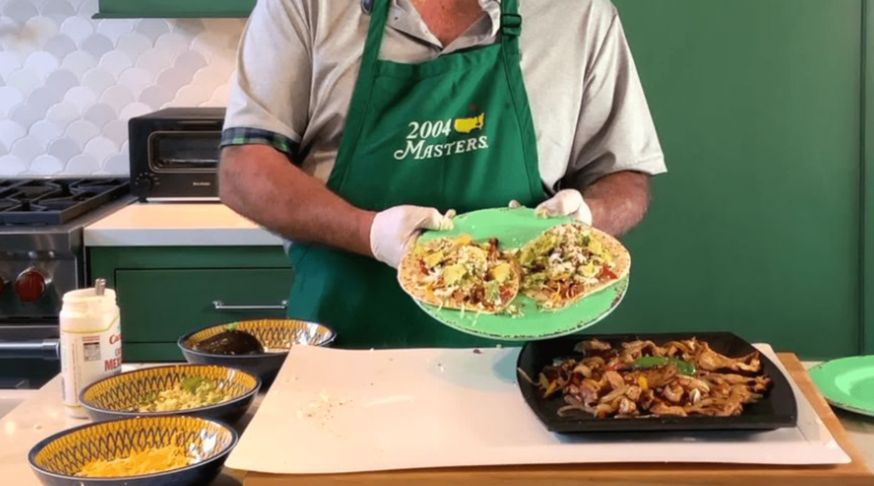 A person in a green apron holds a plate of tacos, surrounded by bowls of ingredients and a skillet of cooked fajita fillings.