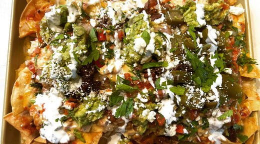 A tray of loaded nachos topped with melted cheese, guacamole, sour cream, jalapeños, fresh herbs, and crumbled cheese.