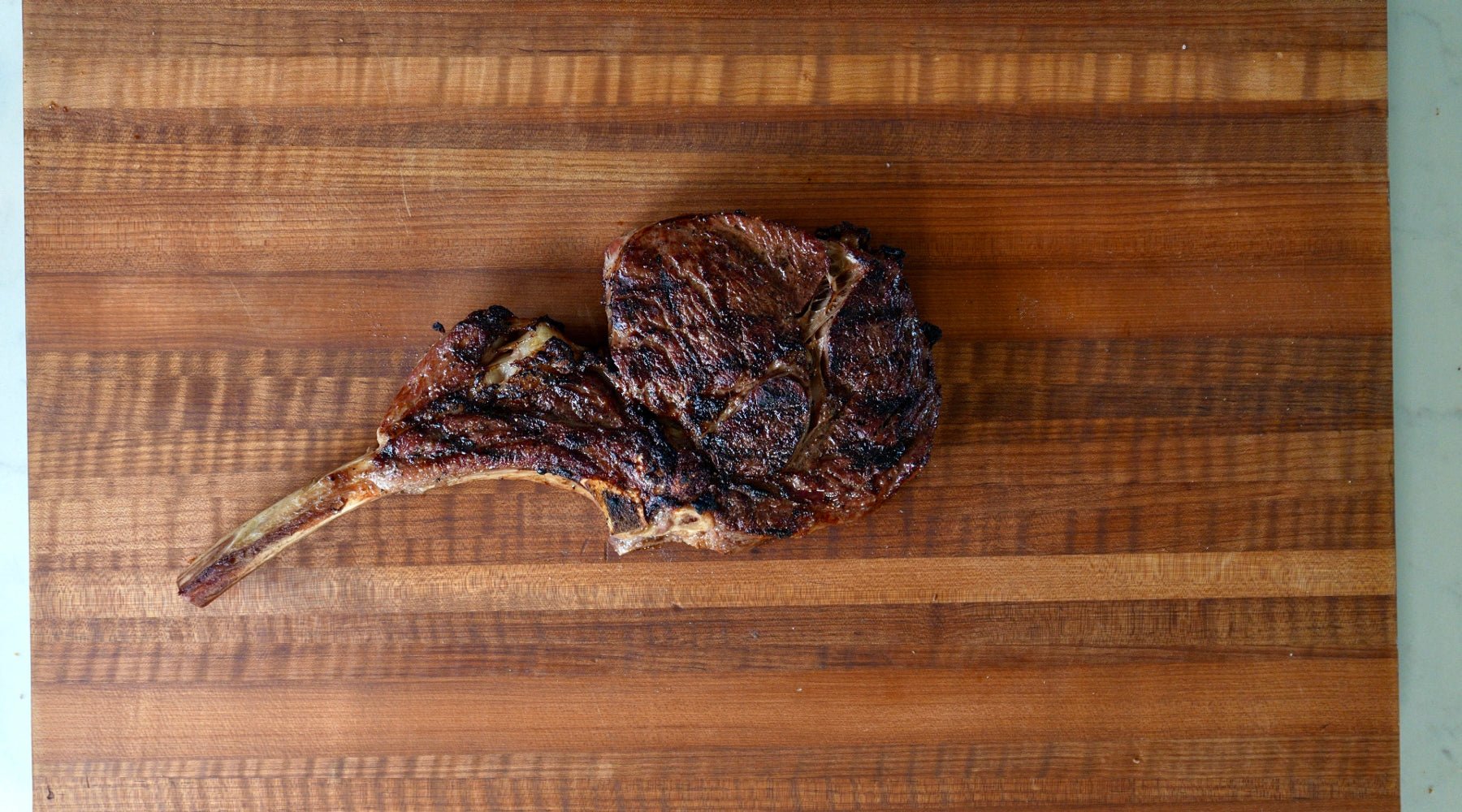 A well-seared tomahawk steak resting on a wooden cutting board. The steak is cooked to a deep brown color with visible char marks, highlighting the bone-in ribeye cut’s marbling and texture.