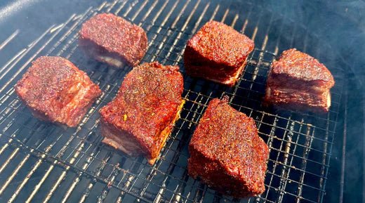 Six short ribs, heavily seasoned with spices, are being grilled on a metal rack. The meat has a deep red color and is cooking over an open flame.