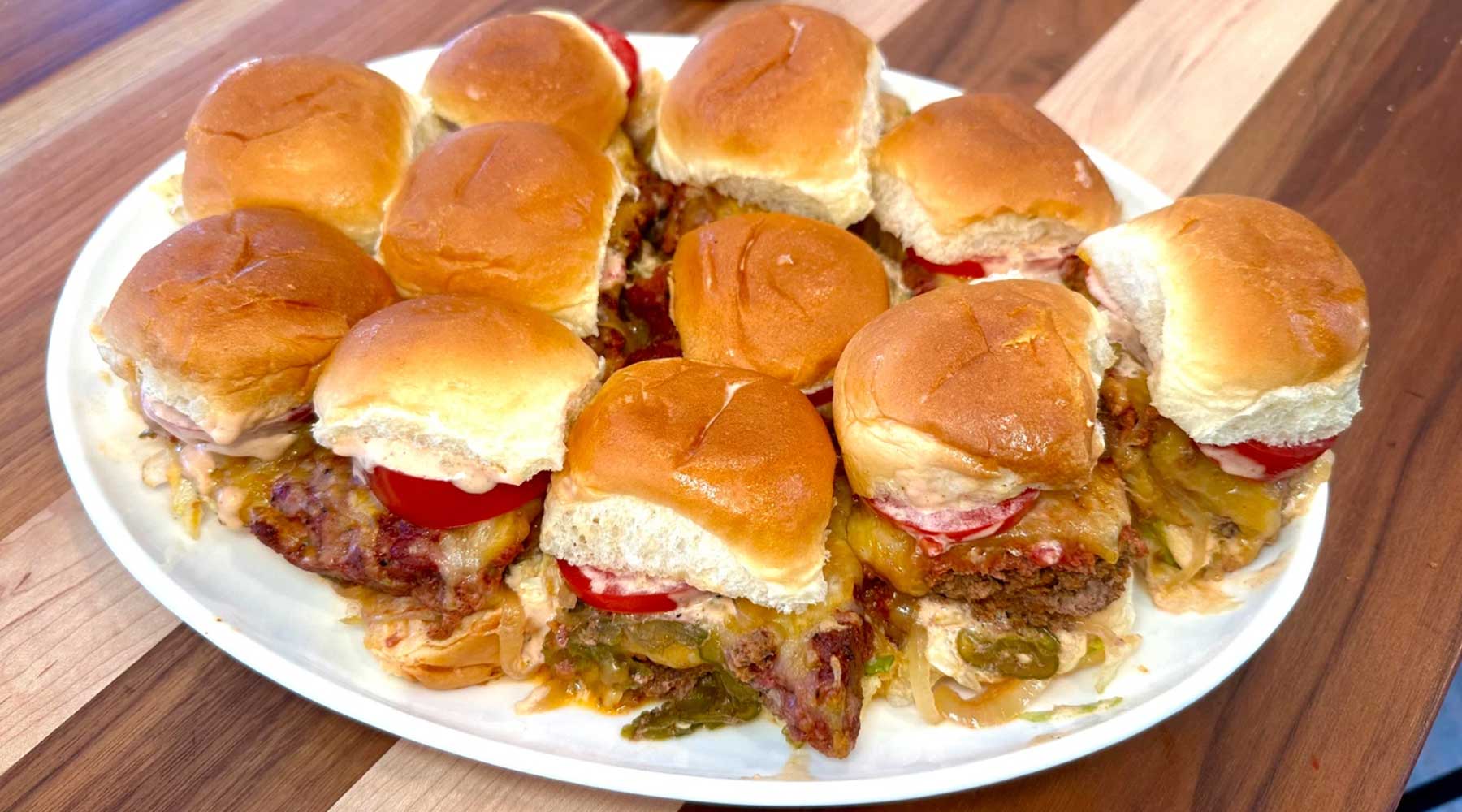 A plate of small sliders on soft, golden buns, filled with melted cheese, sliced tomatoes, and burger patties. The sliders are arranged closely together on a white plate placed on a wooden table.