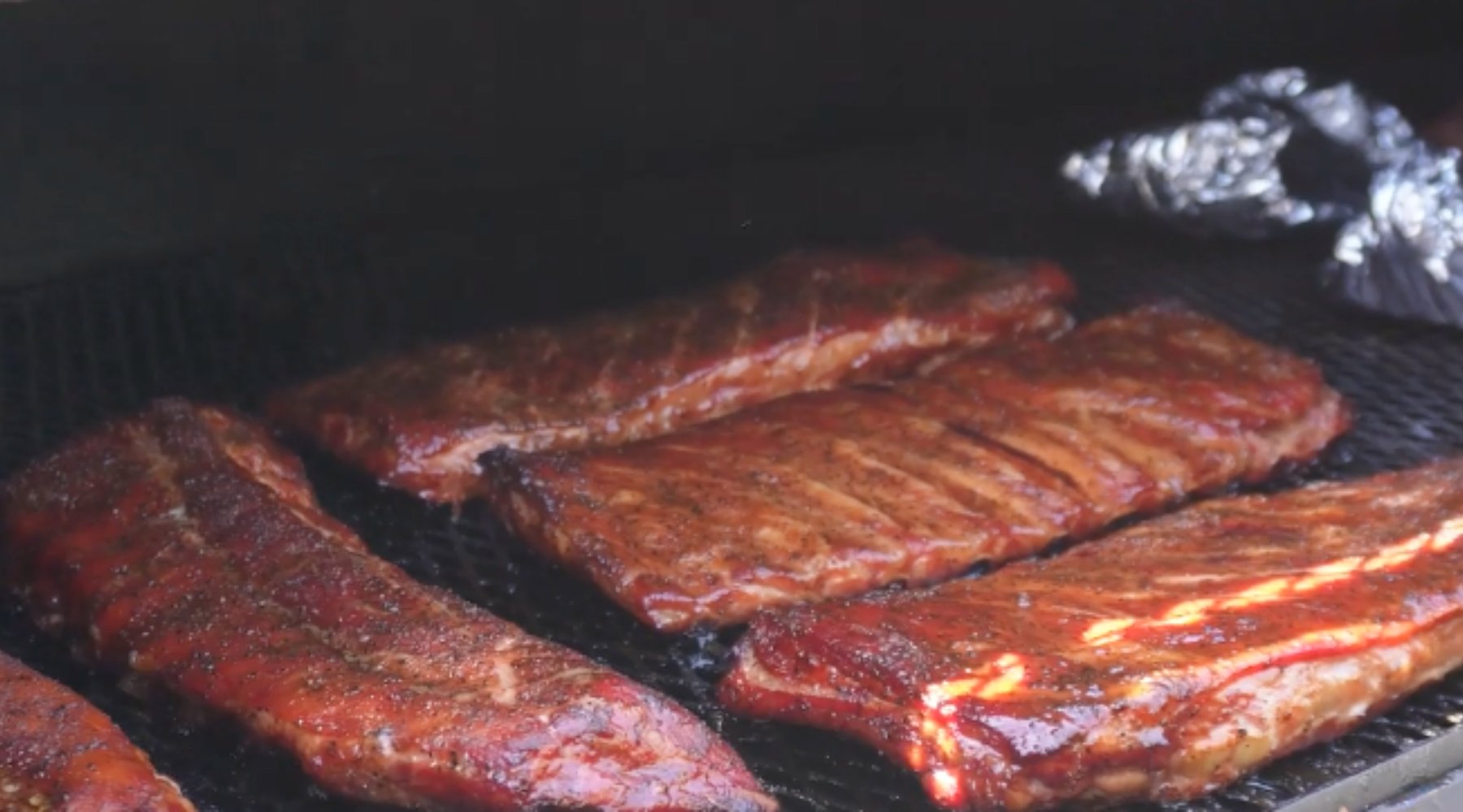 Several racks of pork ribs coated in a rich, reddish-brown glaze are being smoked on a grill, with a light sheen indicating they are almost fully cooked. In the background, there are some foil-wrapped items also being smoked.