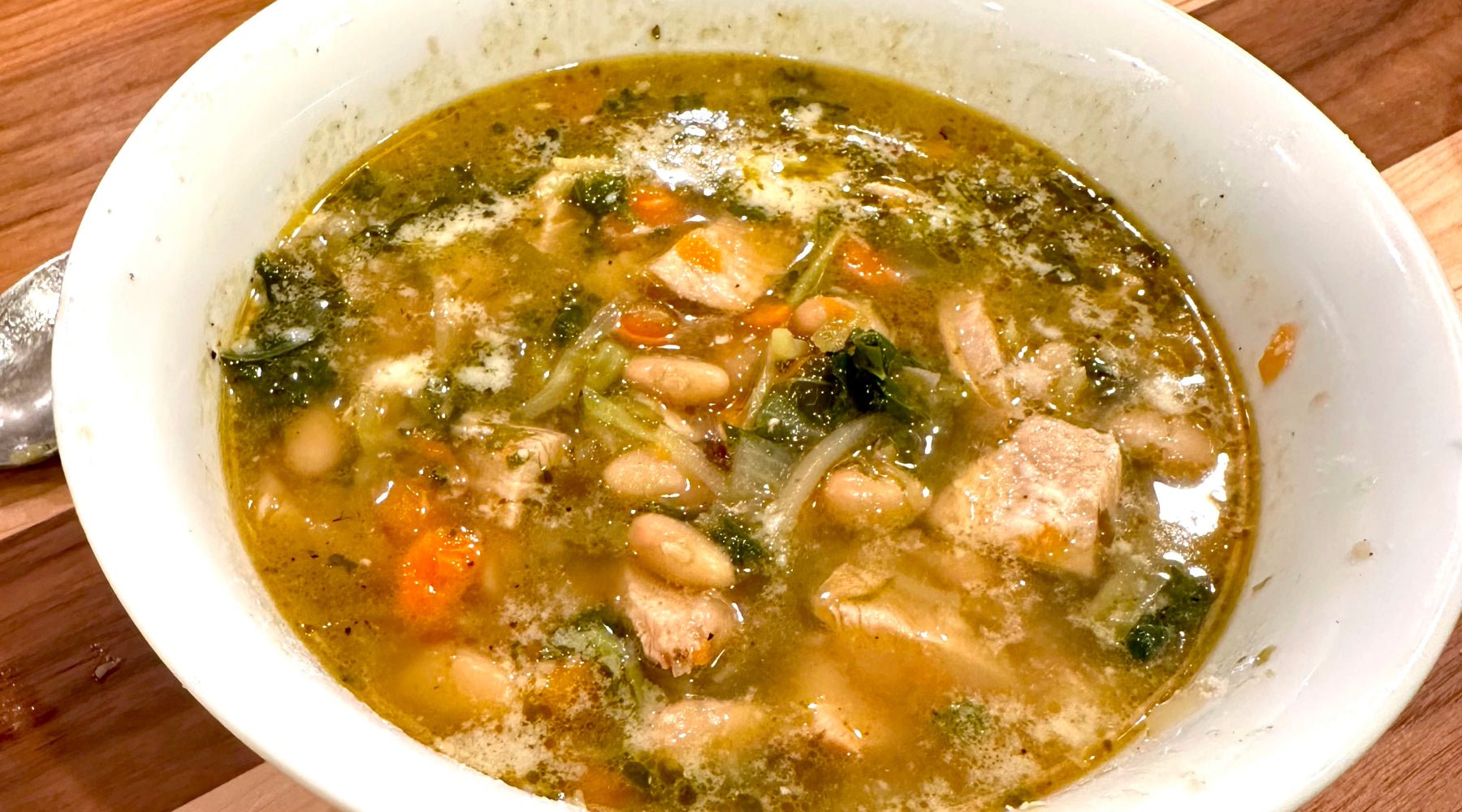 A bowl of hearty soup featuring white beans, chunks of meat, leafy greens, carrots, and onions in a seasoned broth.