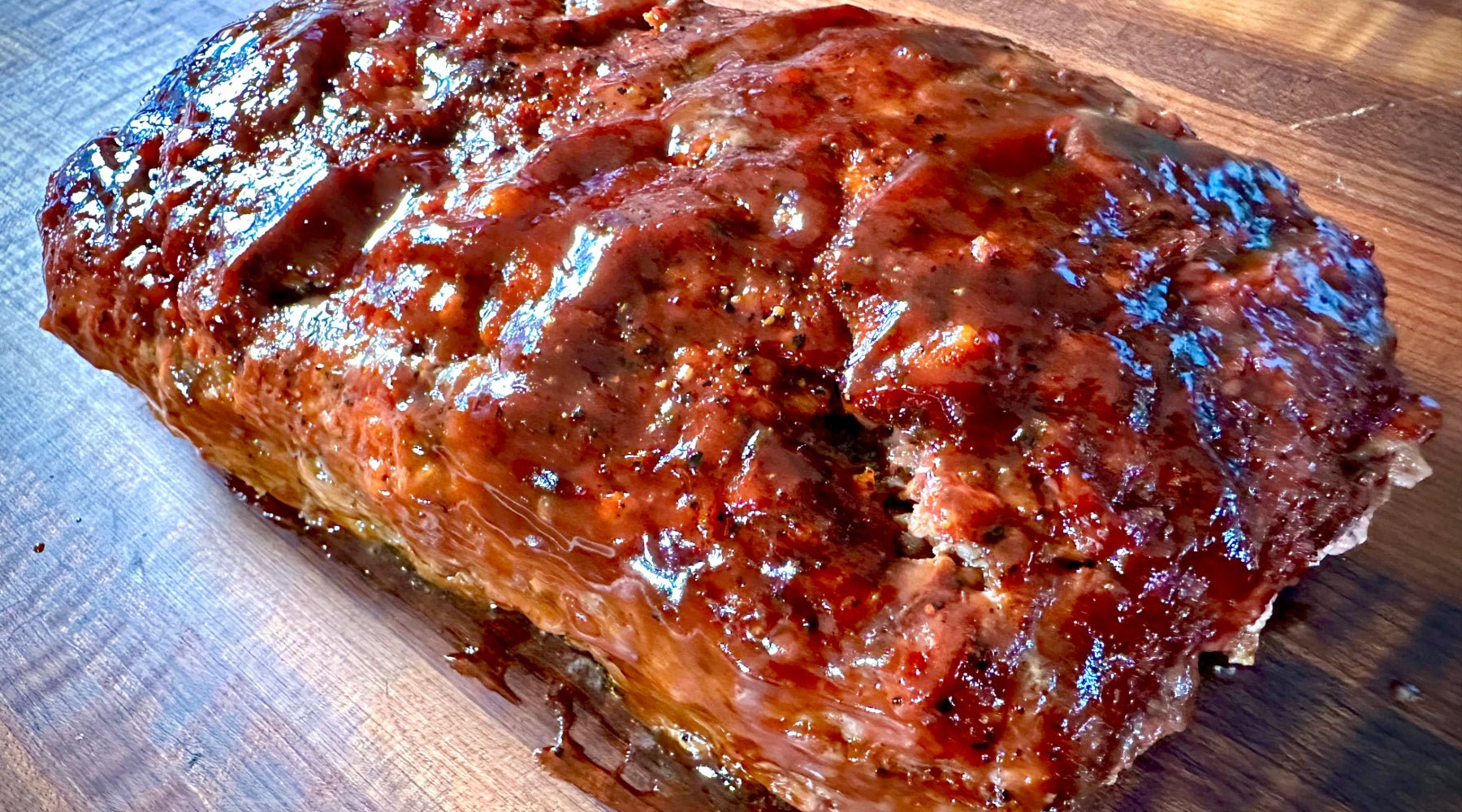 A freshly baked meatloaf coated with a glossy layer of rich, caramelized glaze, resting on a wooden cutting board.